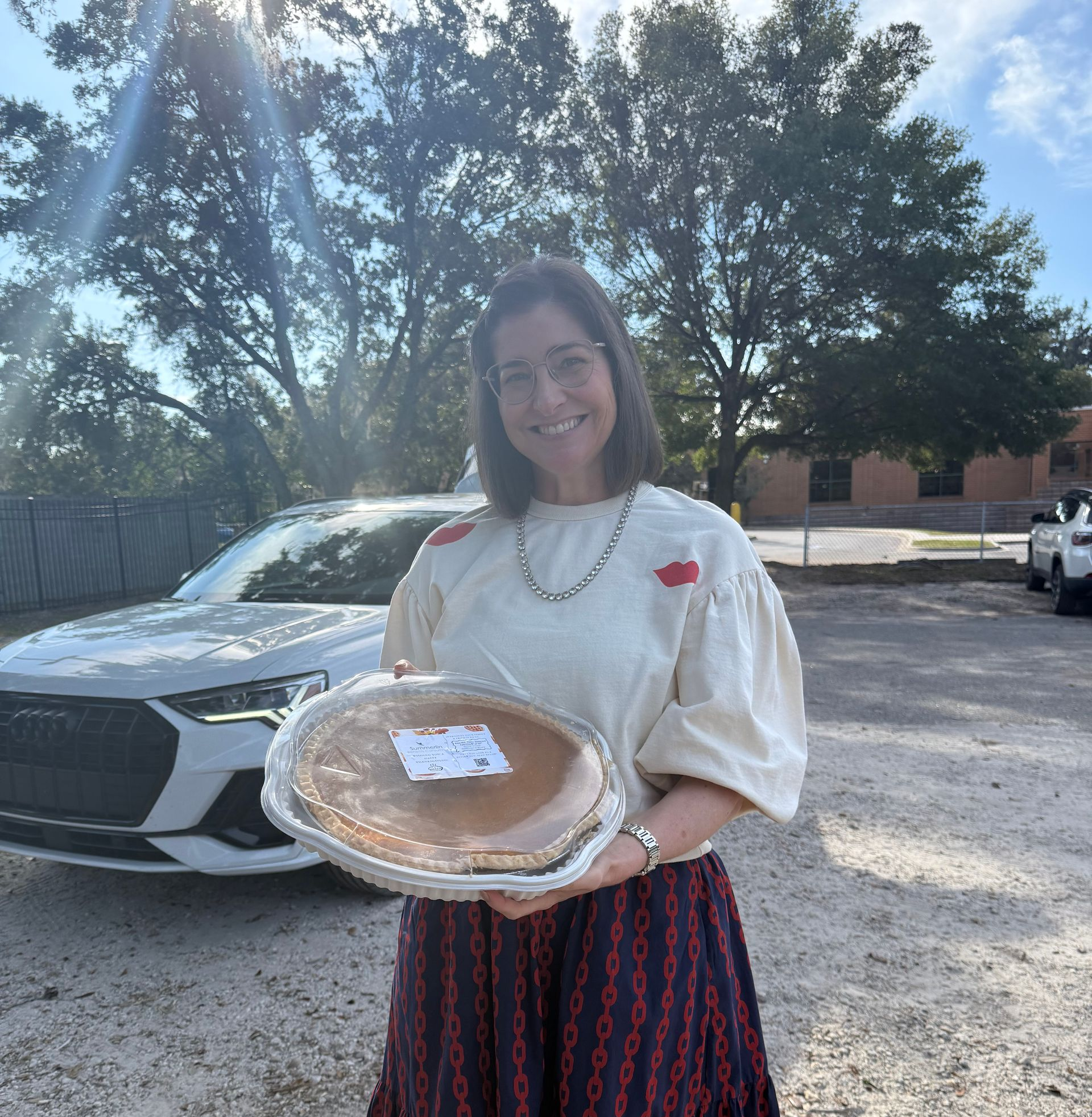 Woman smiling outdoors holding a covered pie tray in front of a parked car.
