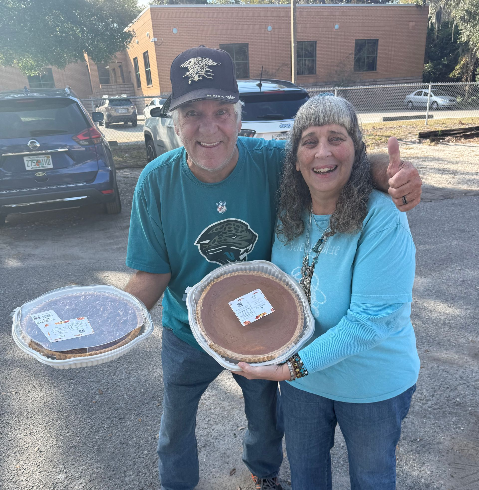 Two smiling people holding baked pies outdoors in a parking lot