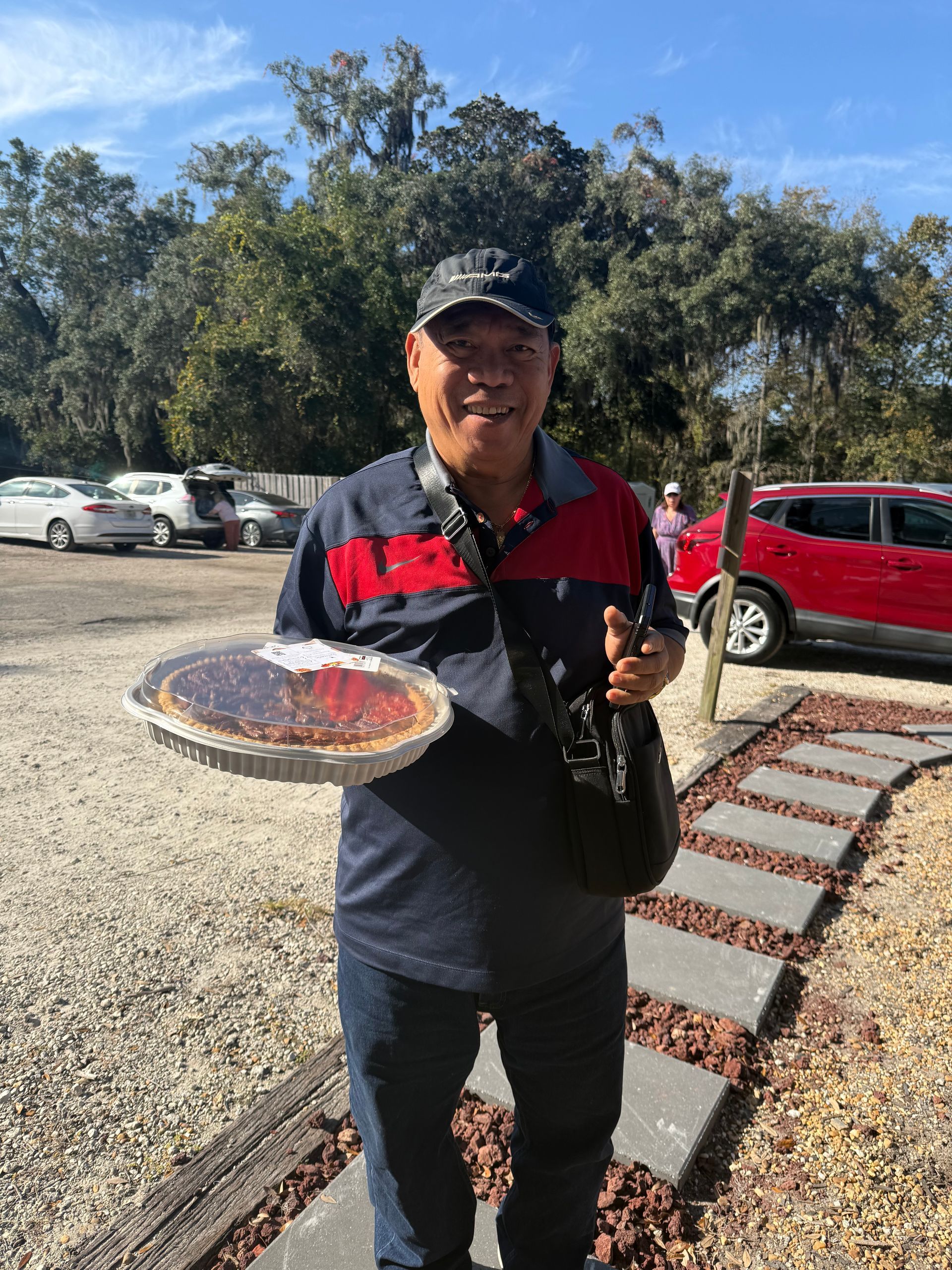 Smiling person holding a pizza tray and bottle beside a red car in a sunny parking lot