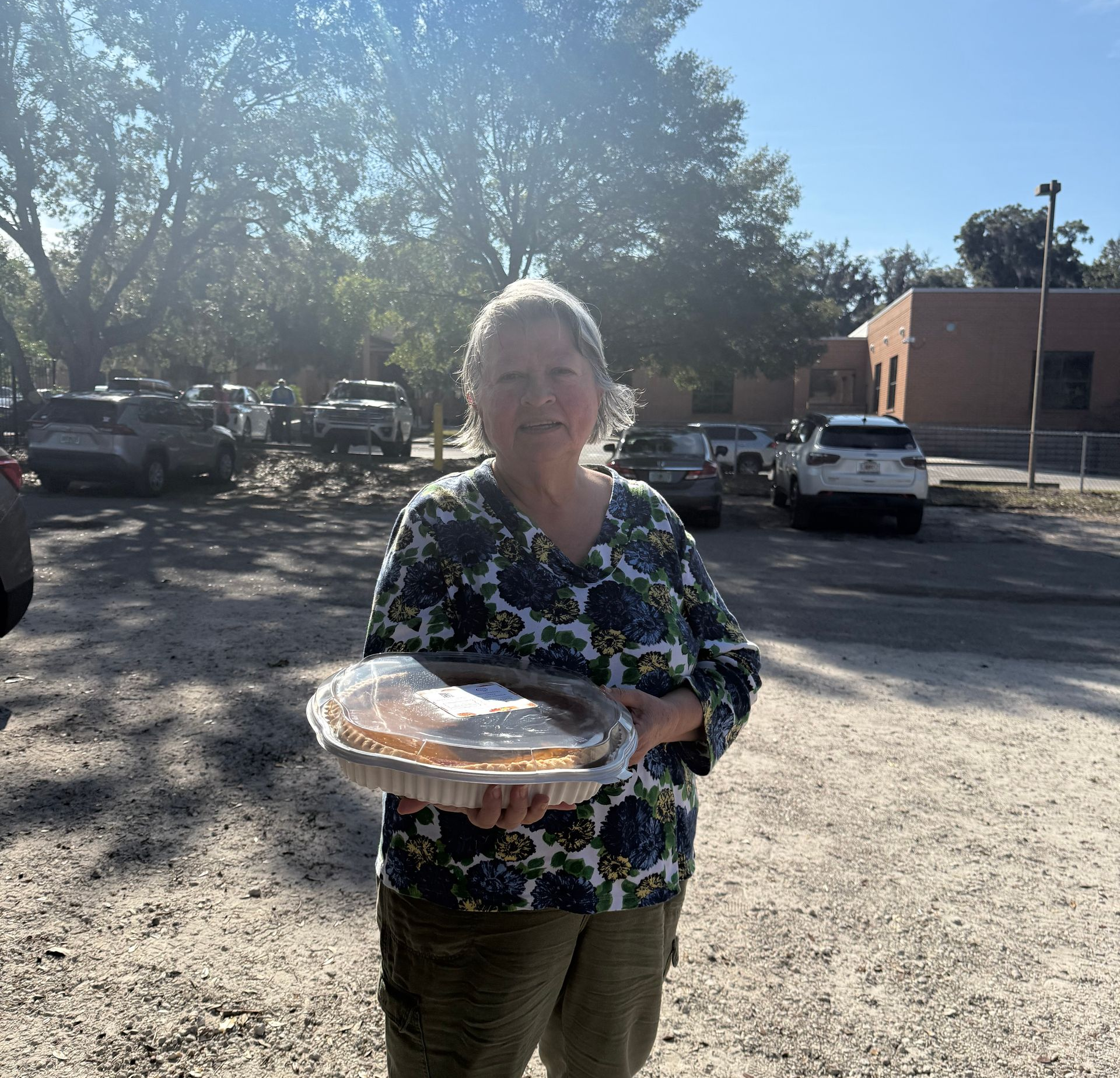 Woman standing outdoors in a parking lot, holding a covered tray in bright sunlight.
