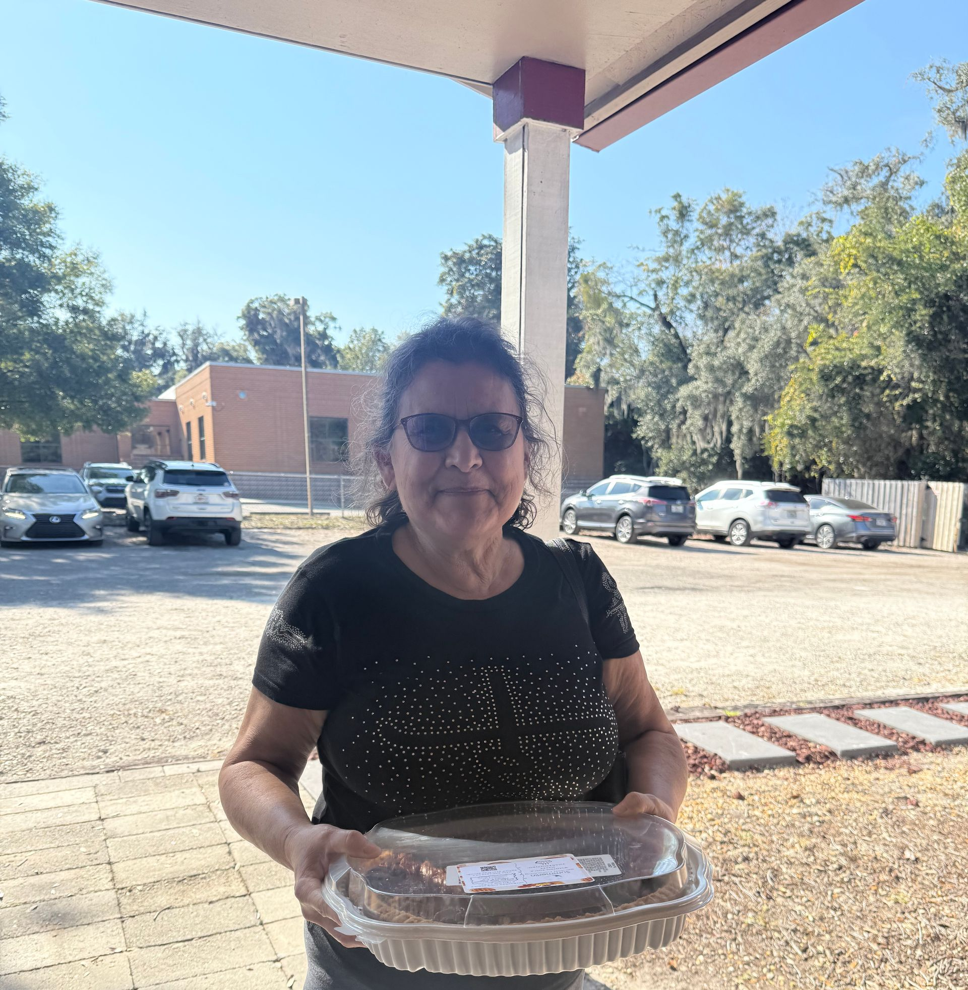 Person in sunglasses holding a foil-covered dish outdoors under a covered patio.