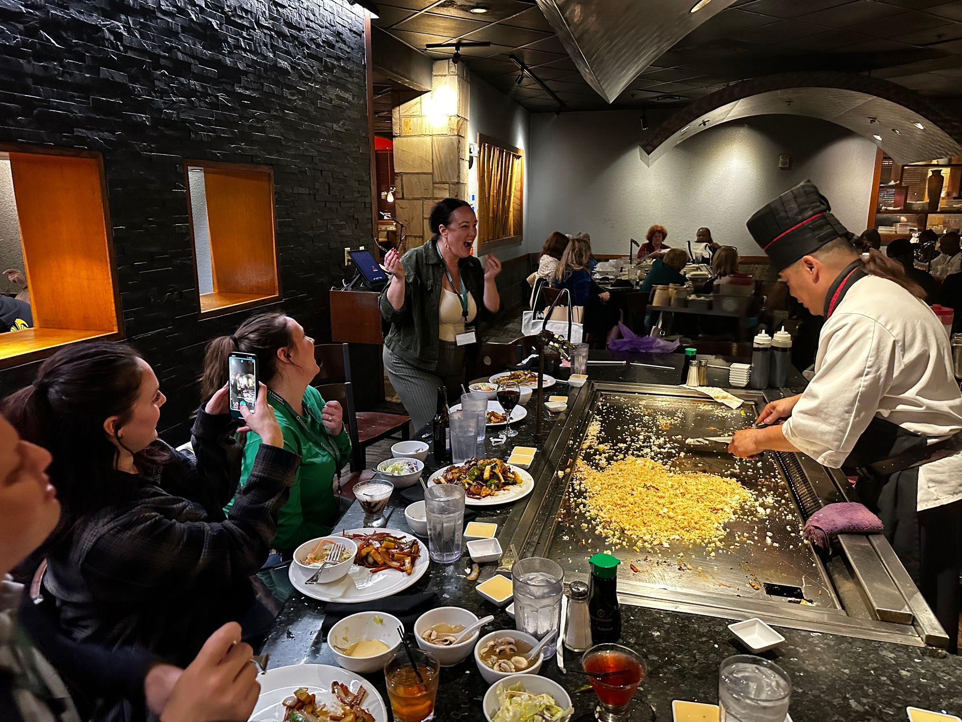 Chef cooking teppanyaki at a lively restaurant table with diners watching and clapping