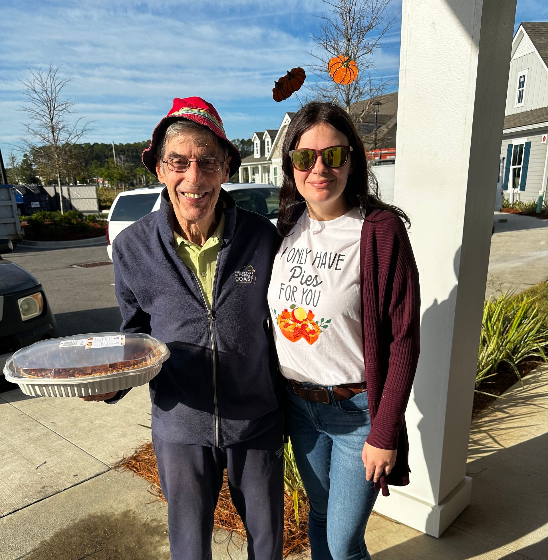 Two people standing outdoors, smiling and holding a pie on a sunny sidewalk