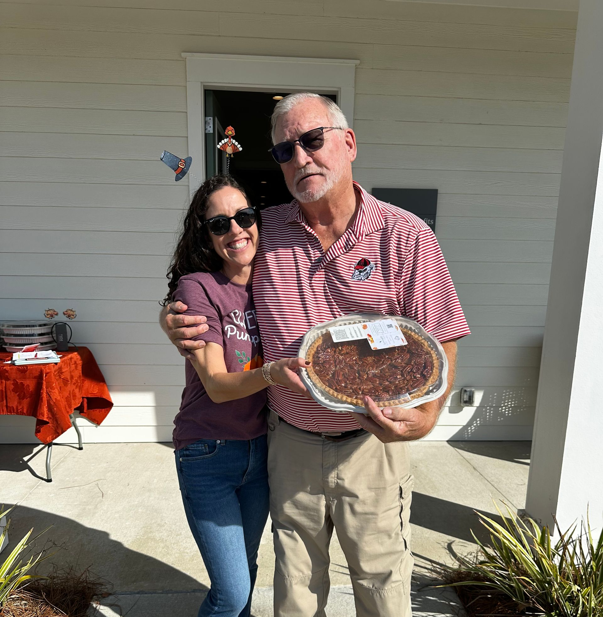 Two people in sunglasses hold a round pie on a sunny porch.