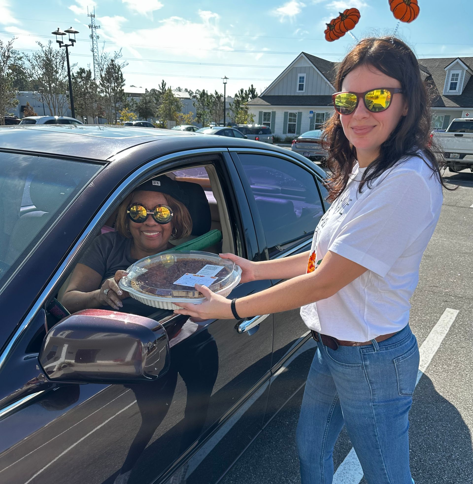 Woman hands a foil tray to a smiling driver at a car window in a sunny parking lot.