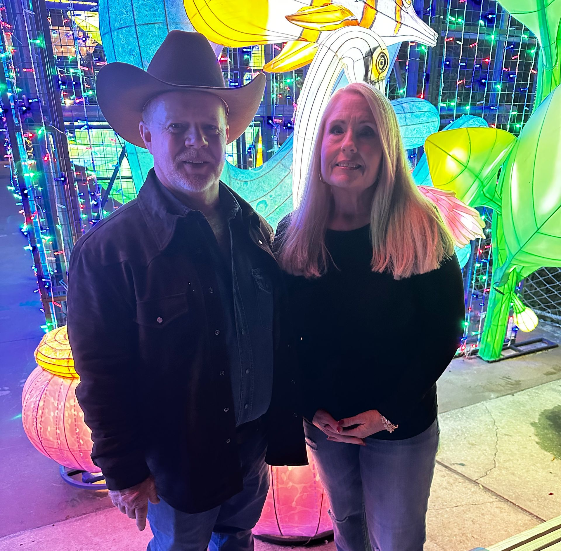 Two people posing in front of a colorful illuminated display, one wearing a black cowboy hat.