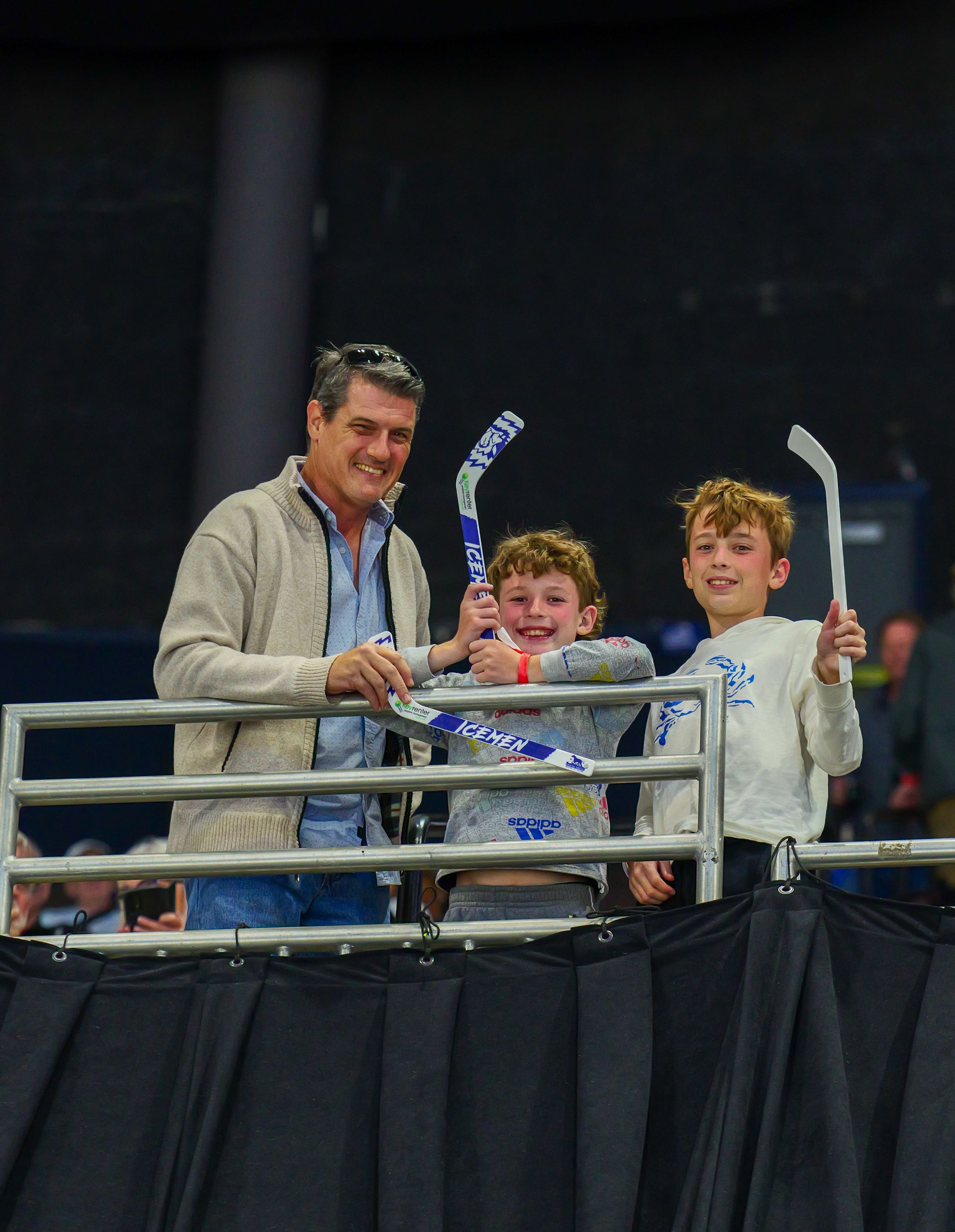 A man and two young boys are holding hockey sticks behind a fence.