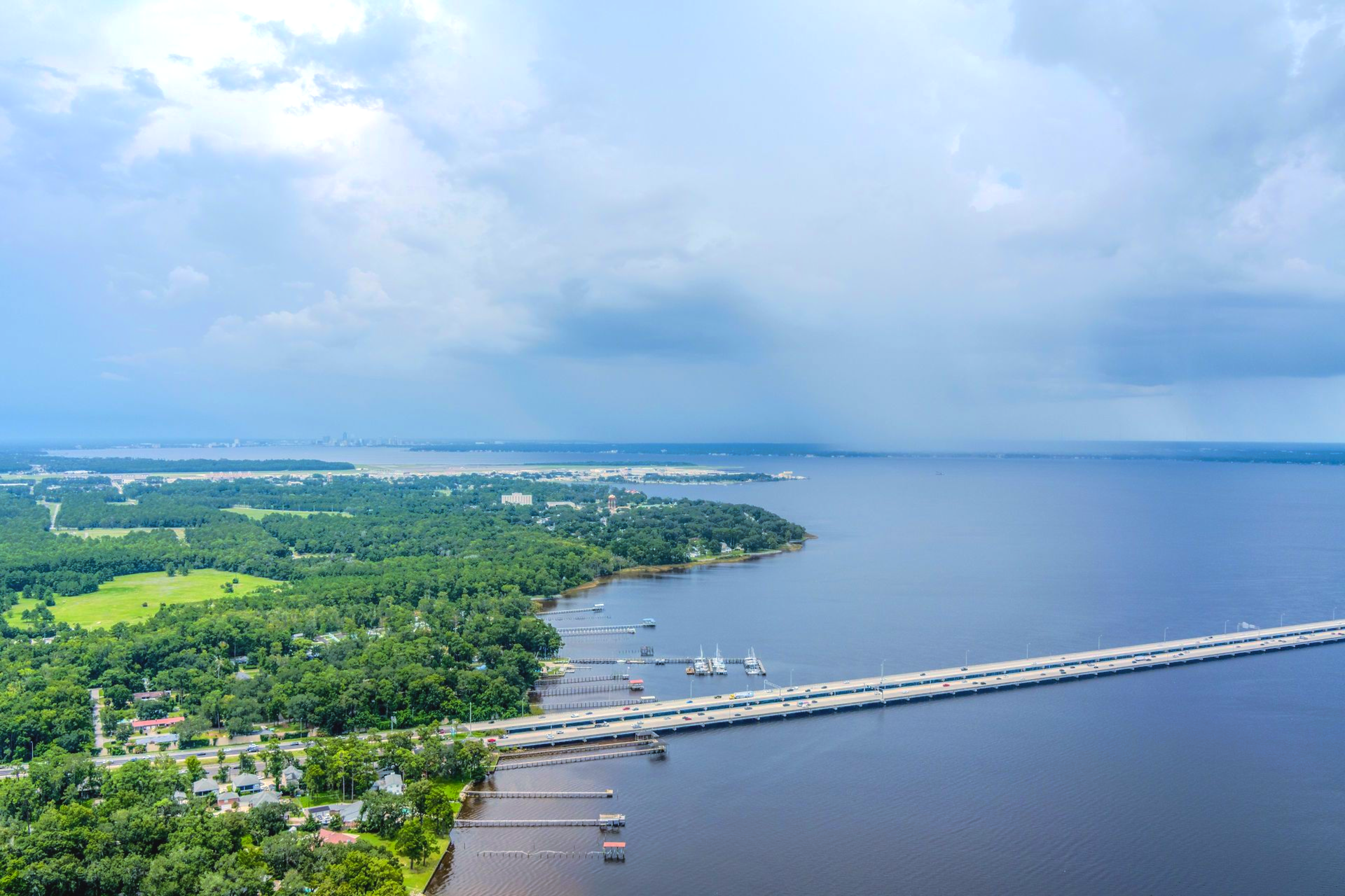 Aerial view of a long bridge over blue water beside a green shoreline under cloudy skies. Orange Park, FL