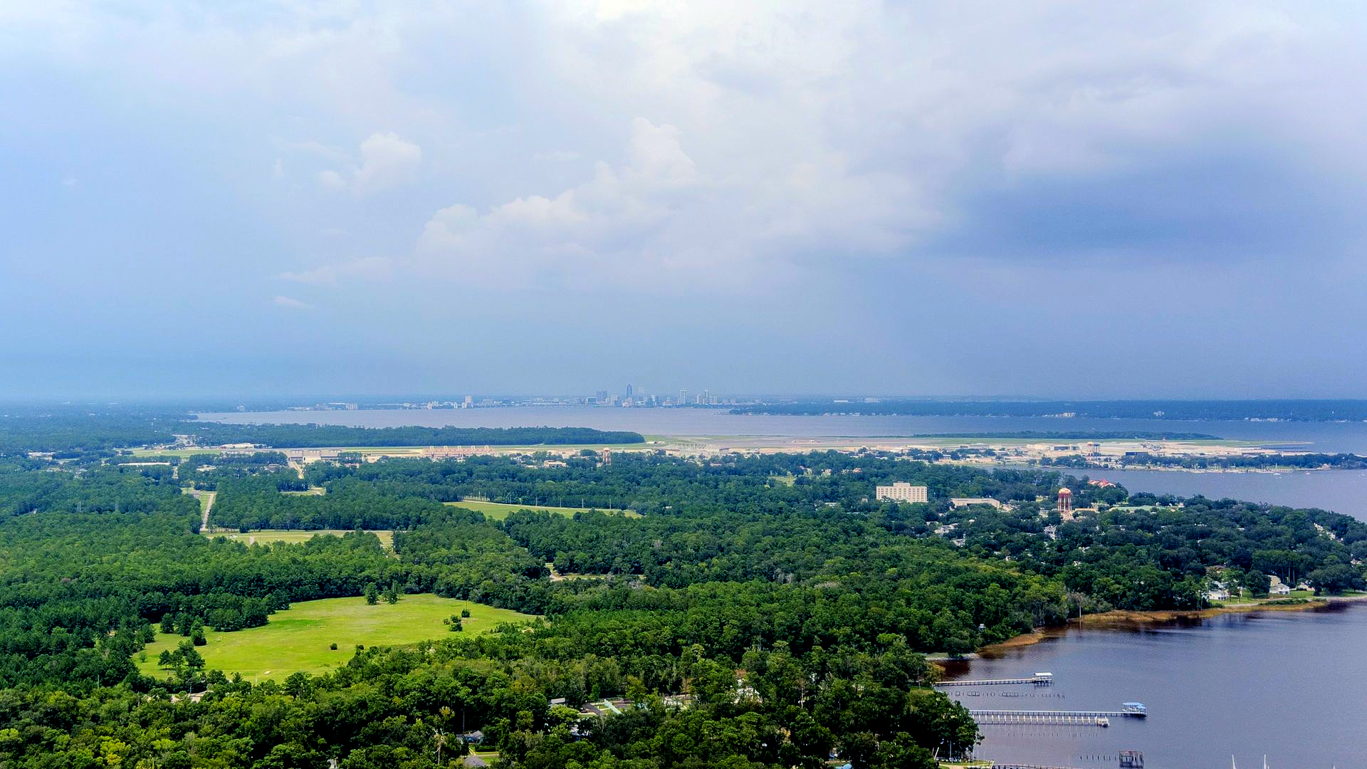 Aerial view of a lush green coastal landscape with a river, shoreline, and distant city under cloudy skies, Orange Park, FL