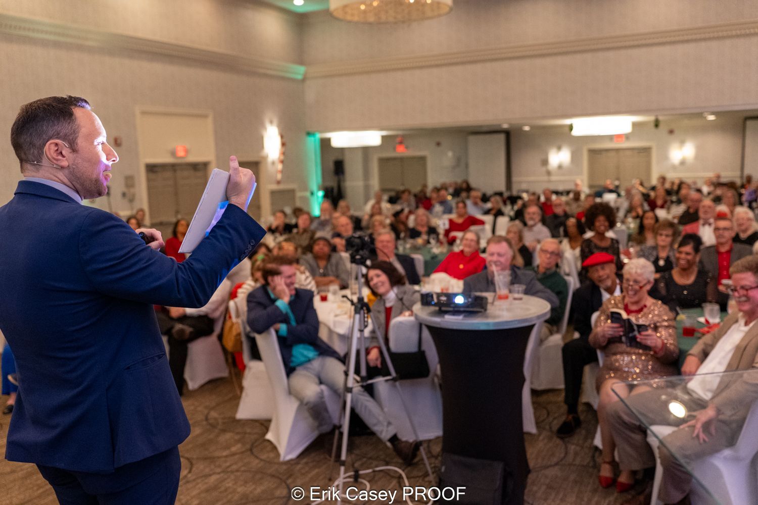 A man in a suit is giving a speech in front of a crowd of people sitting at tables.