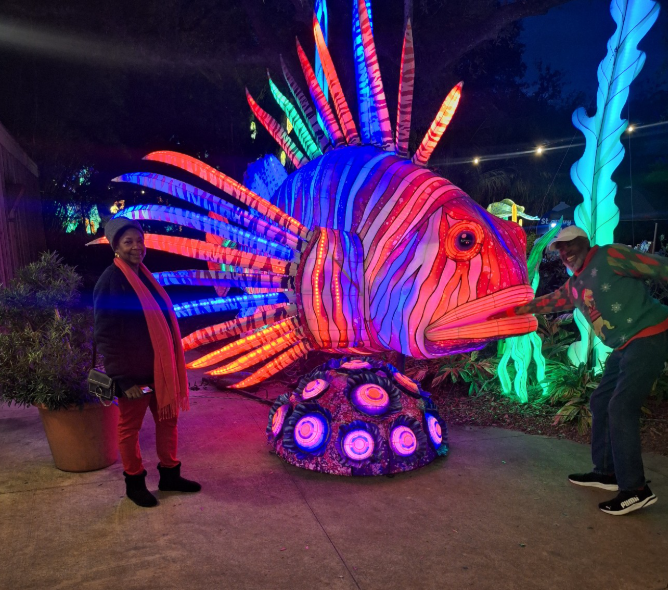 Glowing multicolored fish sculpture at night, with two people standing beside it in a lit outdoor display