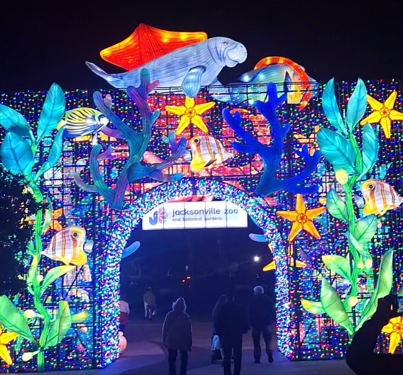 Colorful illuminated marine-themed archway at night with stars, fish, and plants; silhouetted people walk beneath it.