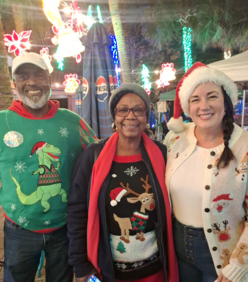 Three smiling people in Christmas sweaters posing outdoors with holiday lights in the background