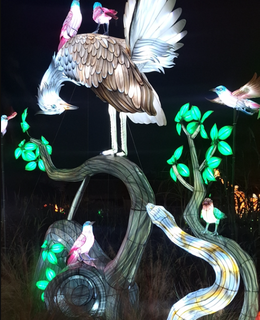 Illuminated bird sculpture with green leaves and a winding stream at night