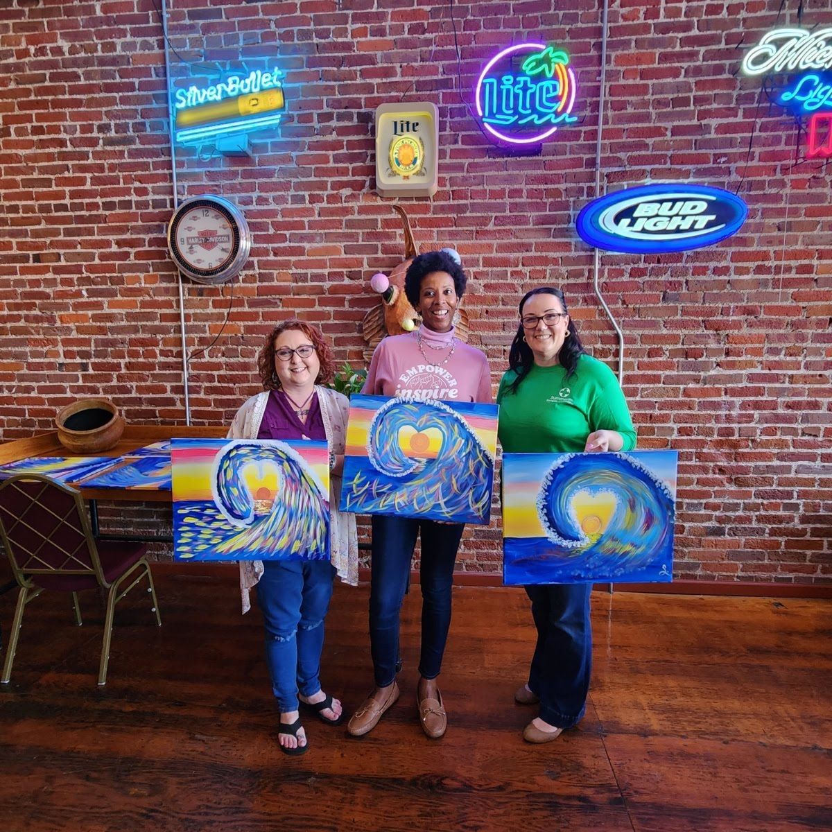 Three people holding colorful paintings in front of a brick wall with neon signs.