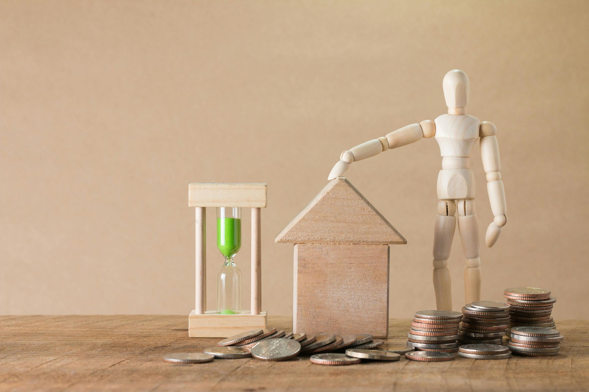 Wooden mannequin beside house model, hourglass, and coin stacks on a table, symbolizing savings and investment