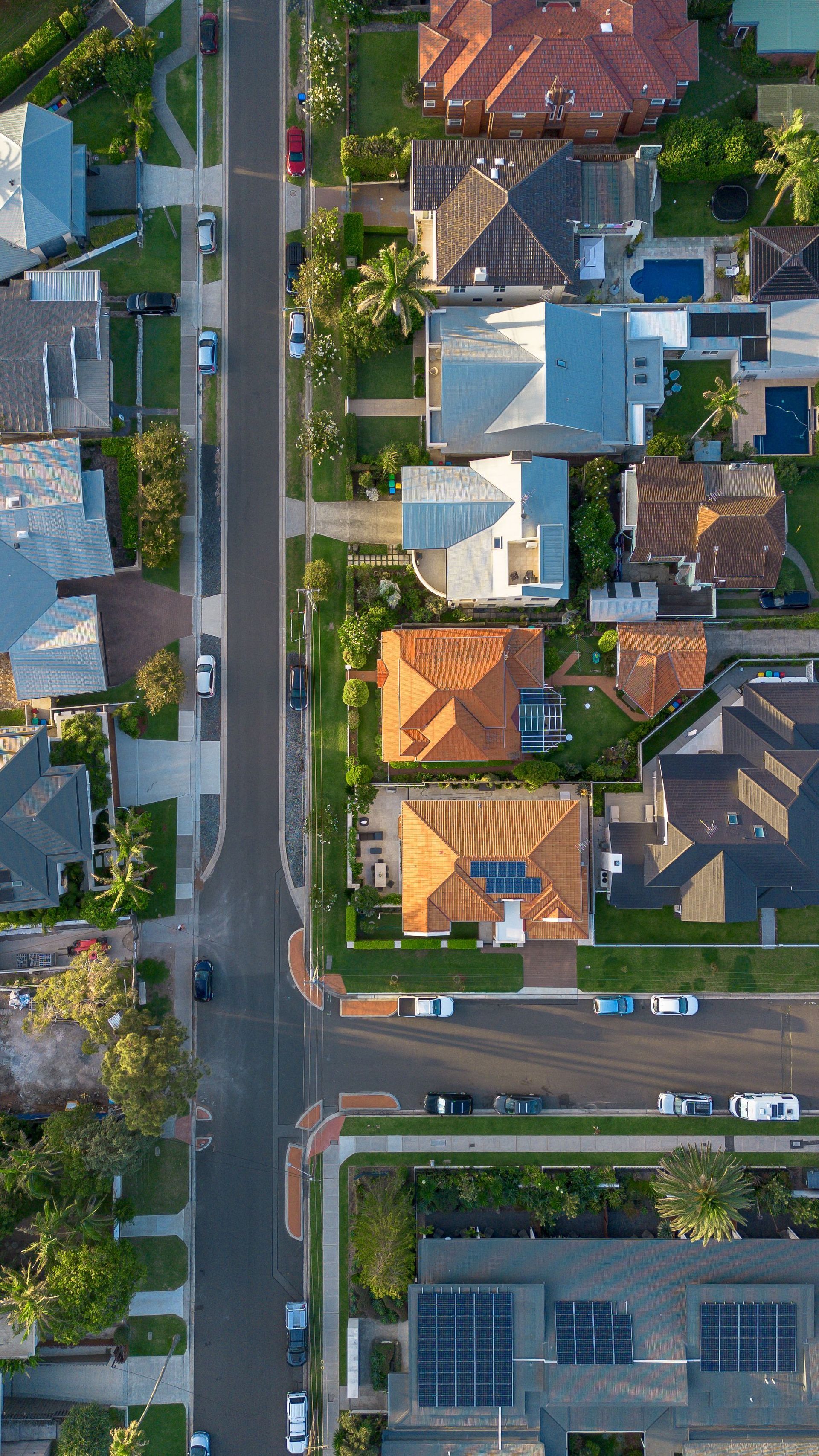 Aerial view of a neighborhood street with parked cars and houses with red, blue, and gray roofs.
