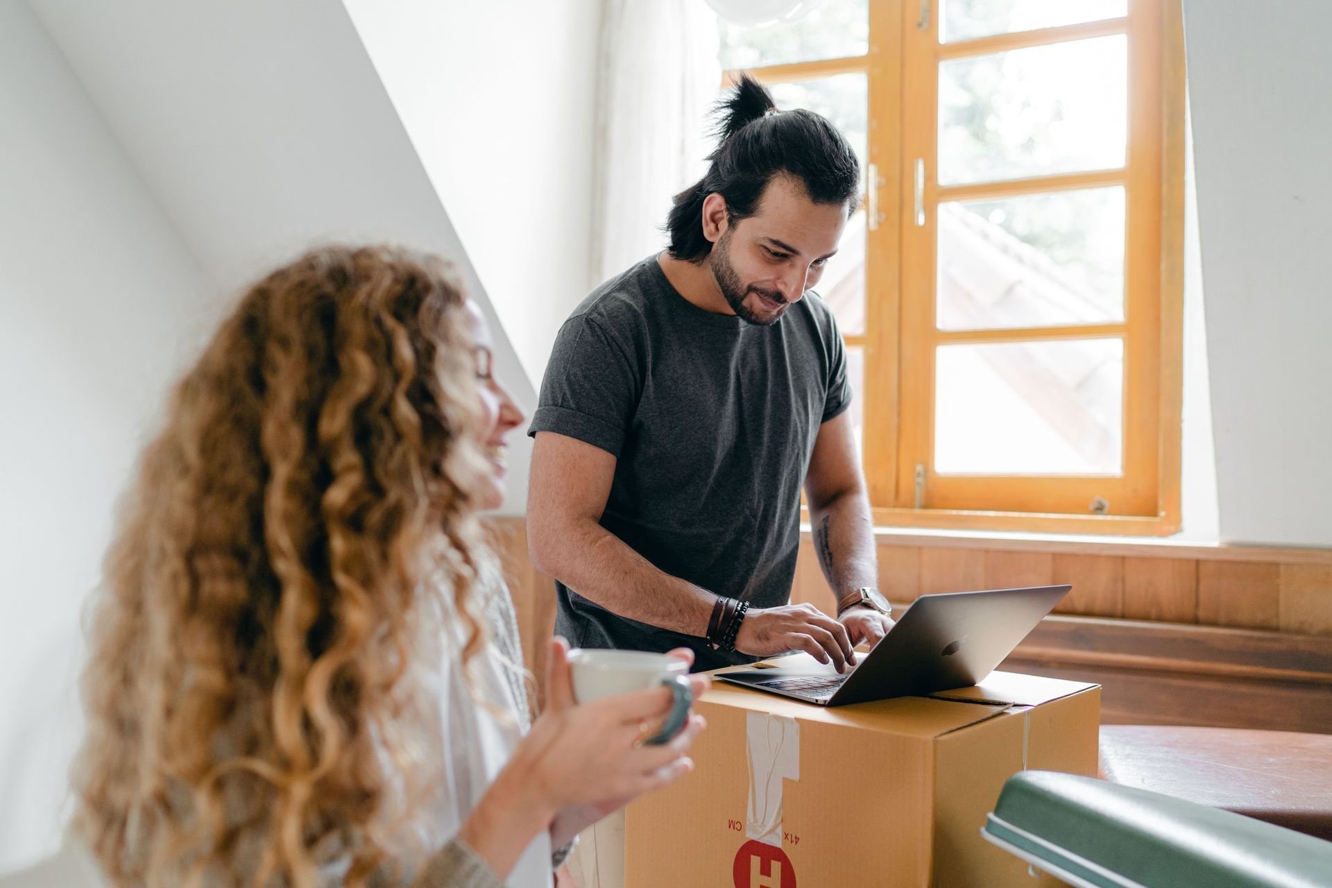 Two people unpacking a box with a laptop in a bright room near a window