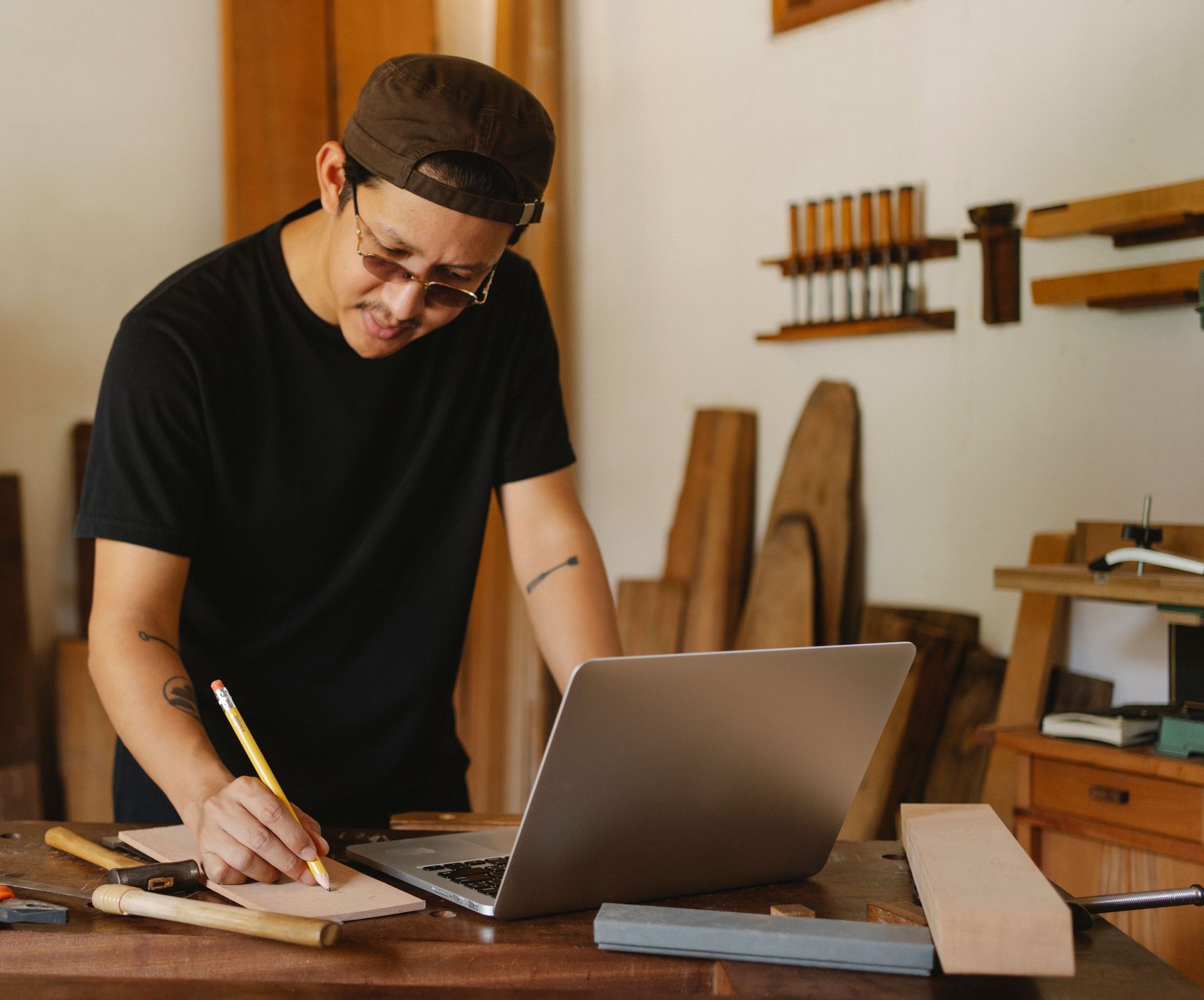 Person sketching a design at a workbench beside a laptop in a woodworking shop