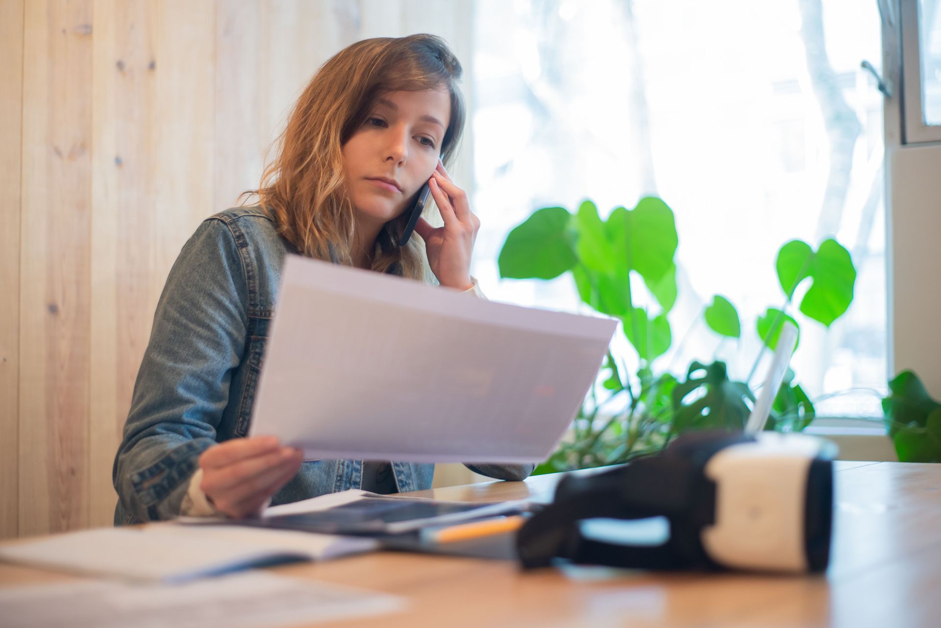 Woman reviewing papers at a desk with a phone by her ear and blurred plant in the background