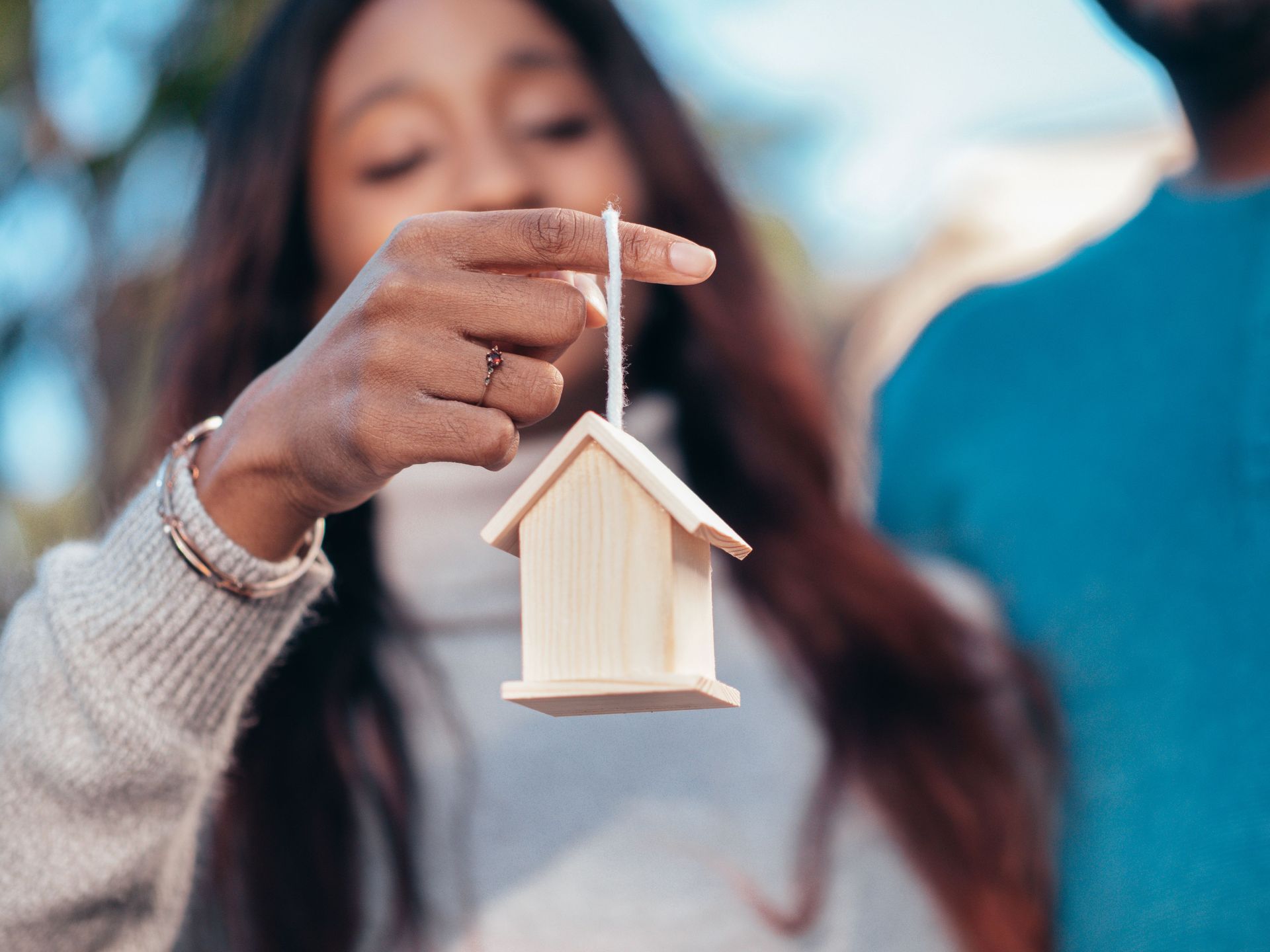 Hand holding a small wooden house ornament on a string, with a blurred background.