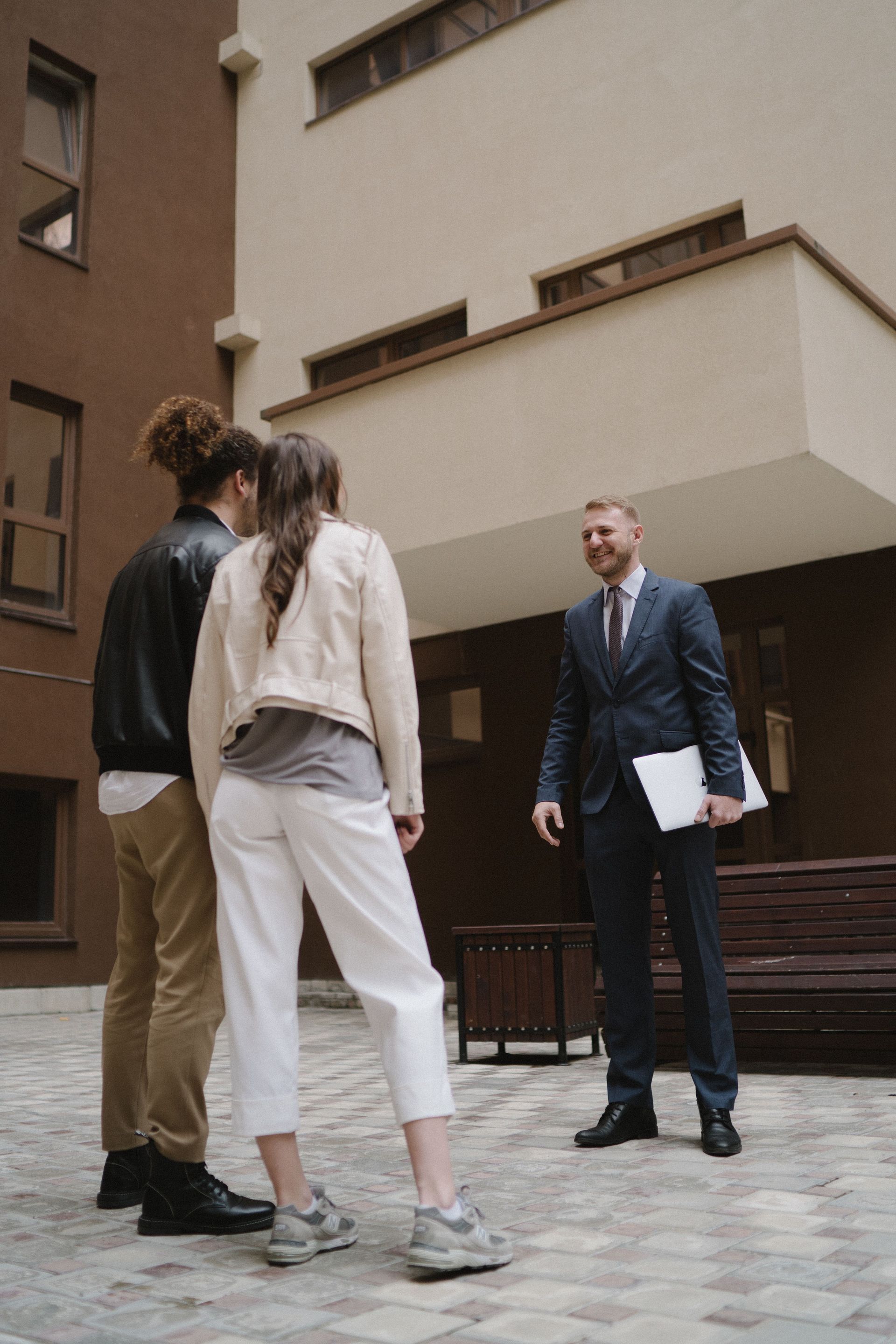 Three people stand in a courtyard, talking to a man in a navy suit holding papers.