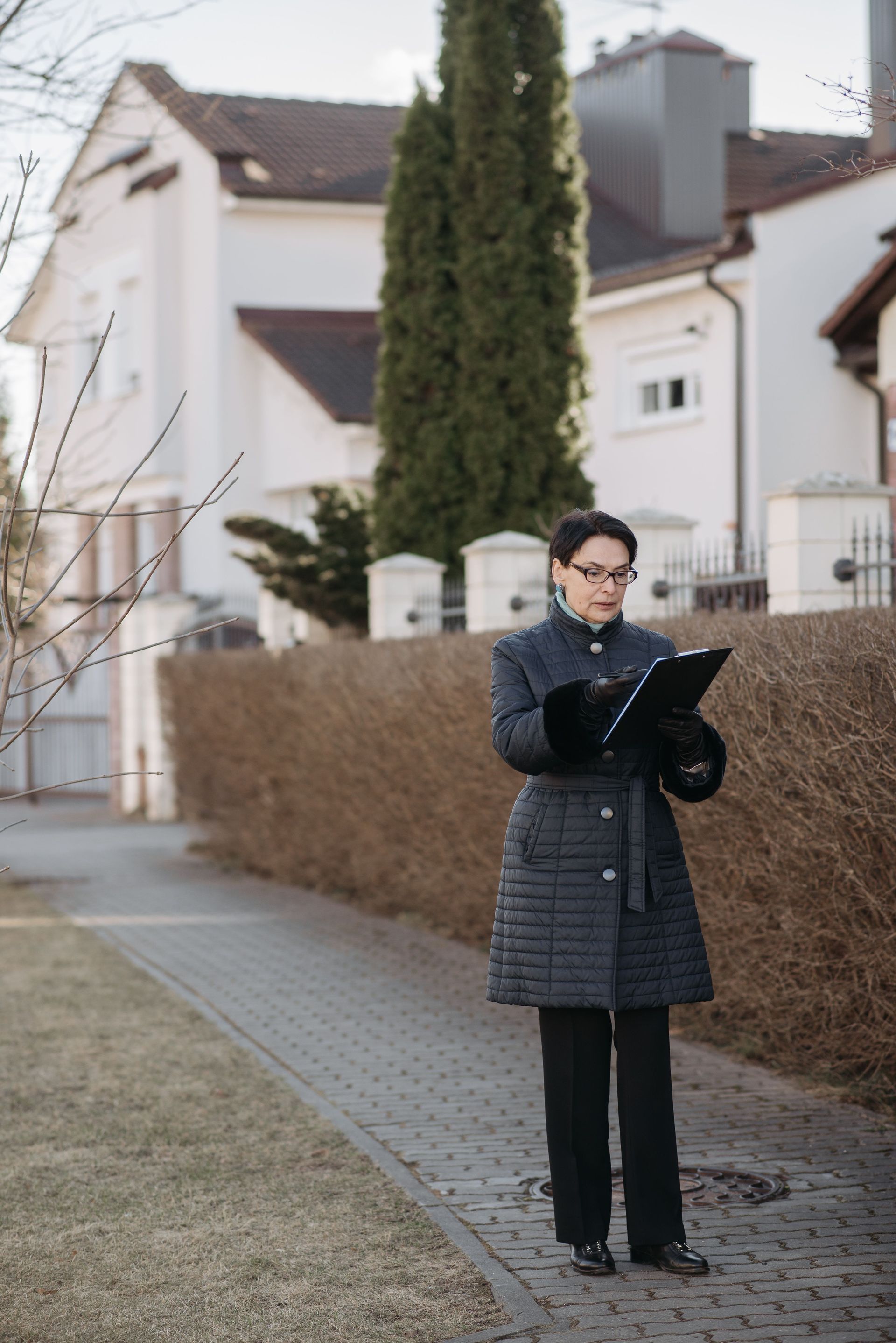 Person in a dark coat standing on a sidewalk, looking at a phone in a residential neighborhood.