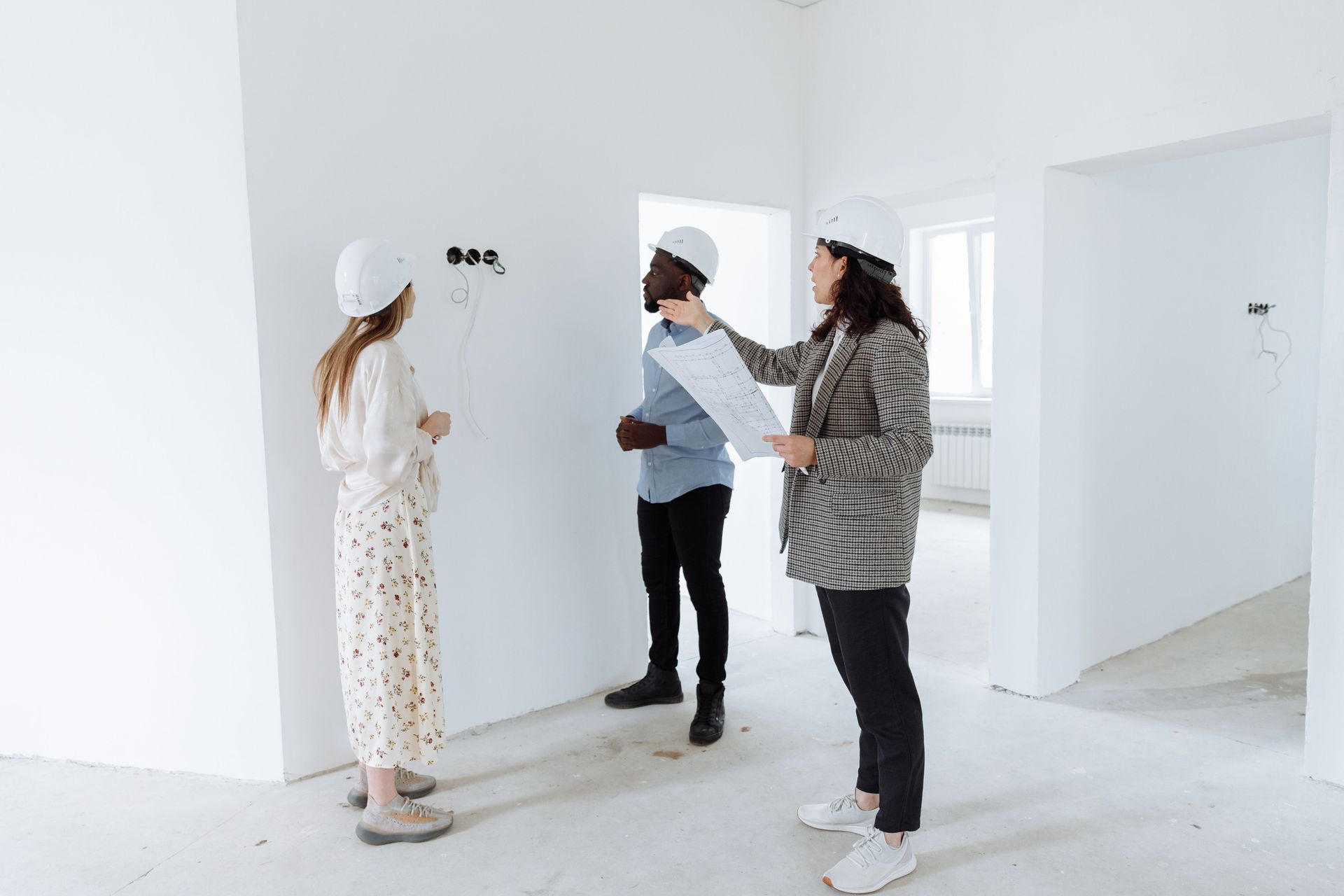 Three people in a bright empty room, discussing a wall while one points at it.