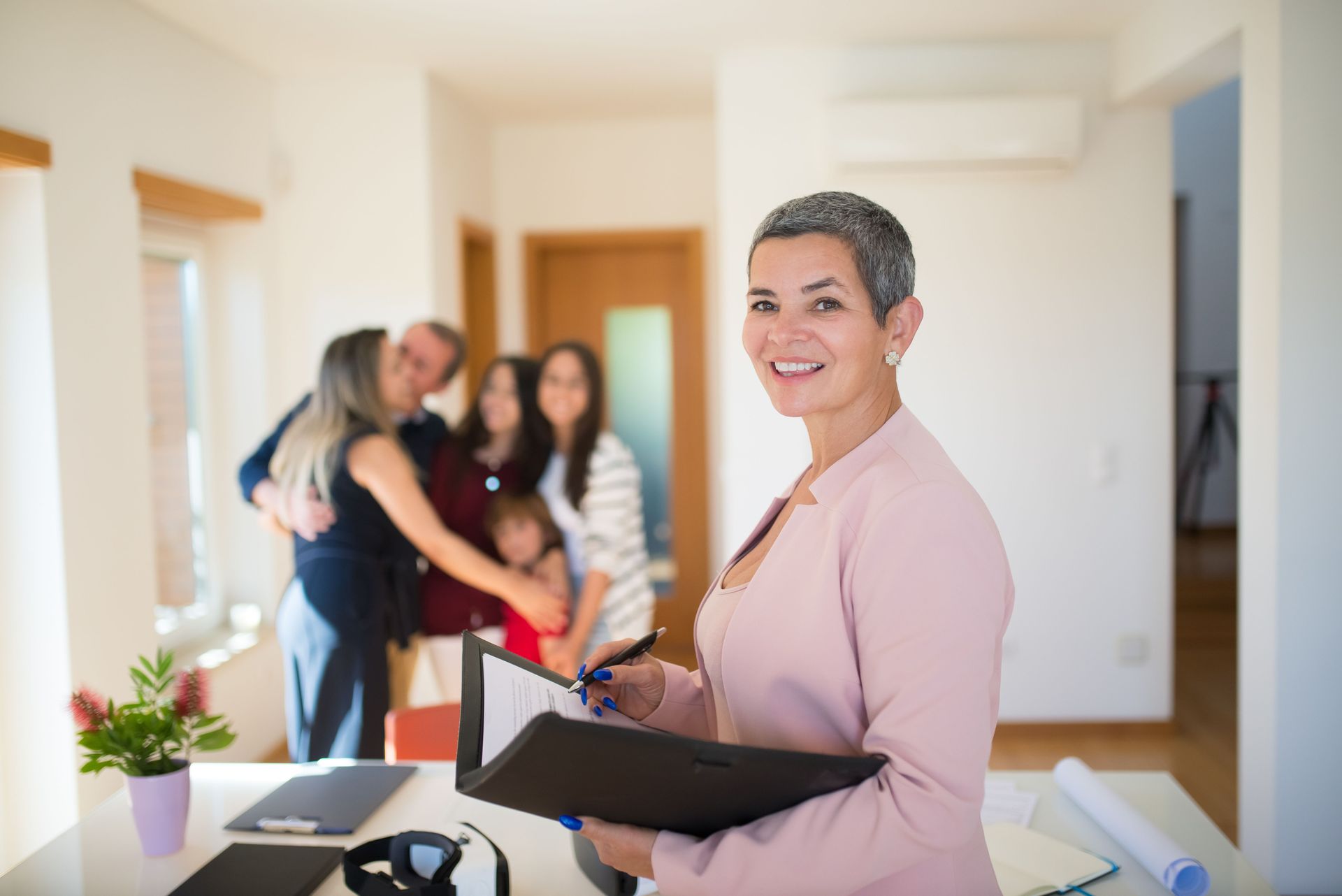 Smiling woman holding a folder in a bright hallway while a blurred group chats in the background