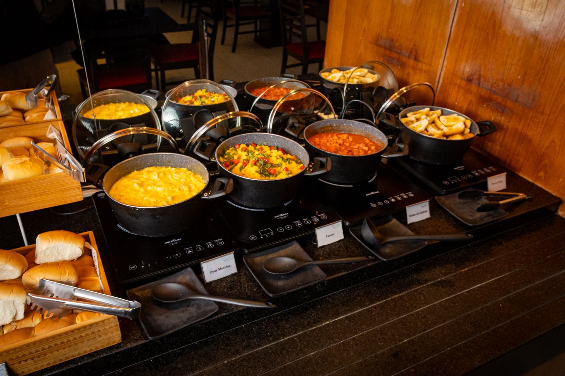 Buffet com diversos pratos em panelas pretas sobre uma superfície aquecida; pãezinhos à esquerda.