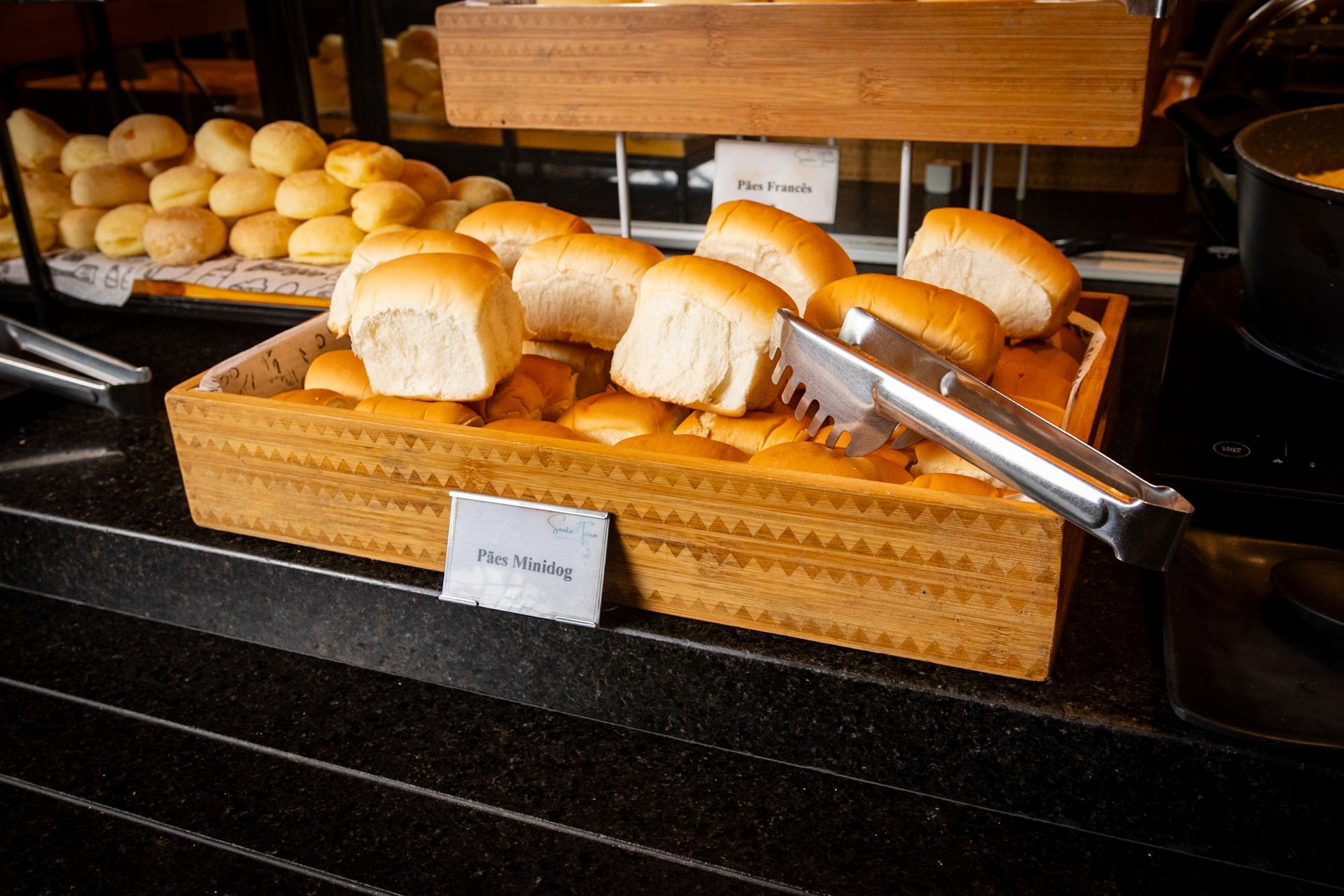 Pãezinhos em uma cesta de madeira com pinças, ao lado de pãezinhos menores e uma placa que diz 