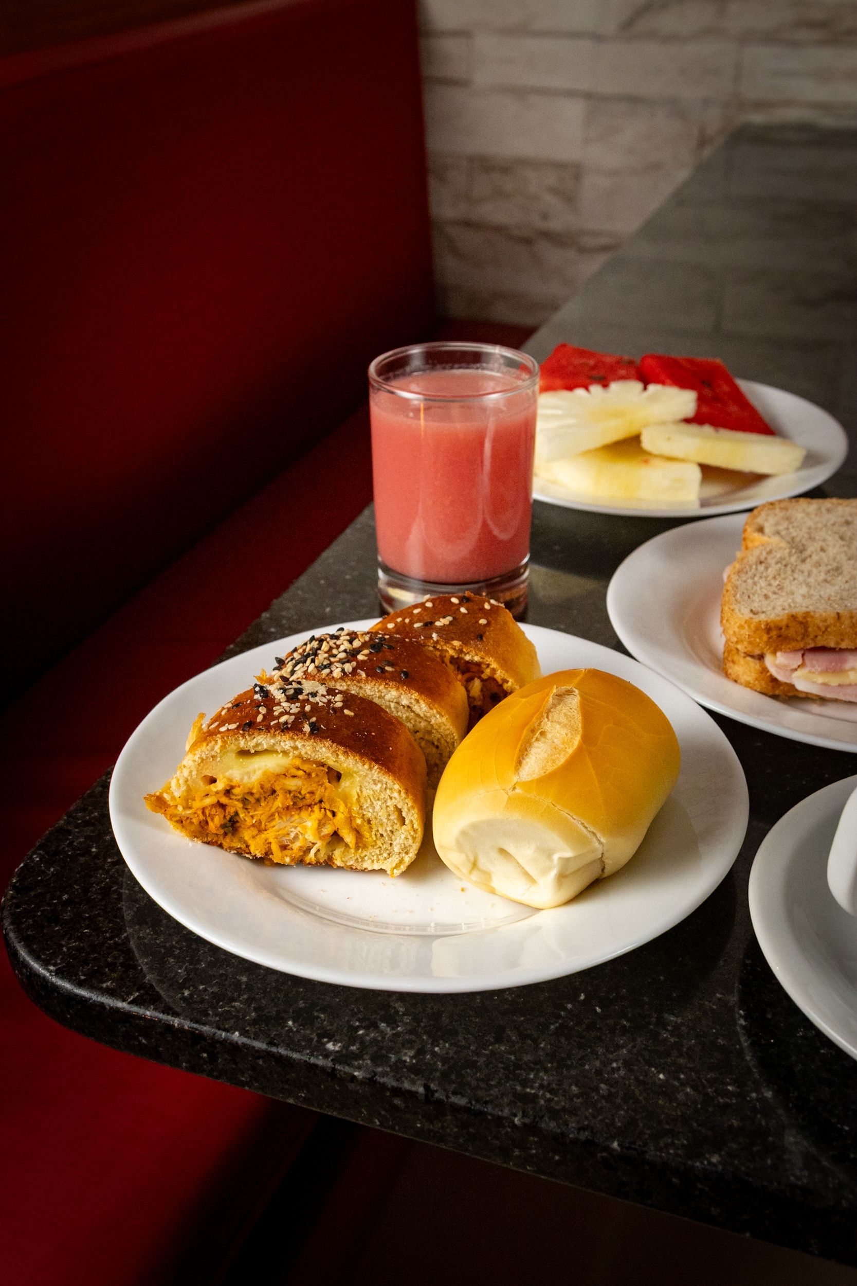 Café da manhã variado: doces, pãezinhos, sanduíche, frutas, suco sobre uma mesa, ao lado de assentos vermelhos.