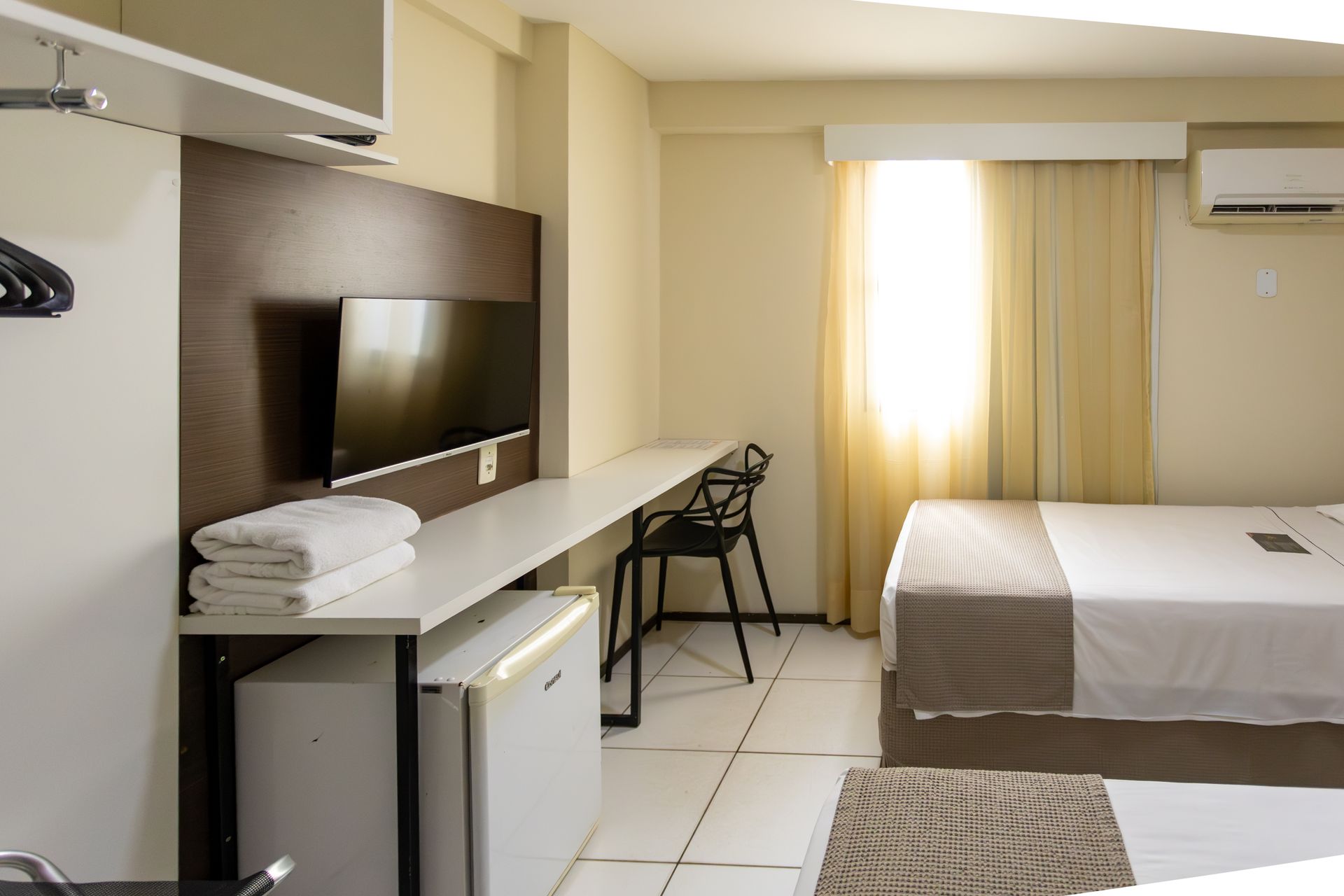 Quarto de hotel com cama, escrivaninha, TV, frigobar e janela.