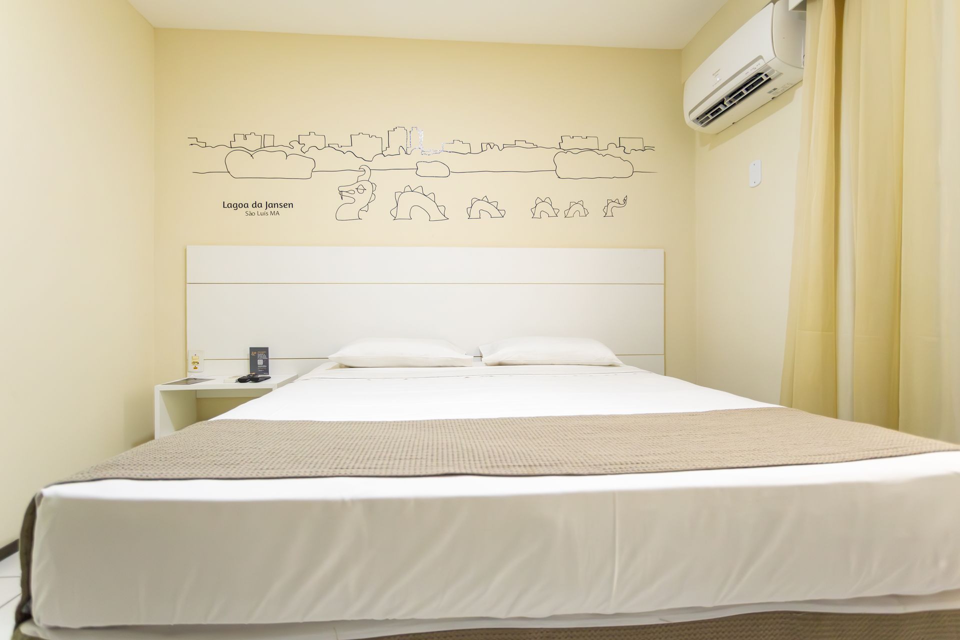 Quarto com cama branca, paredes bege e aparelho de ar condicionado.