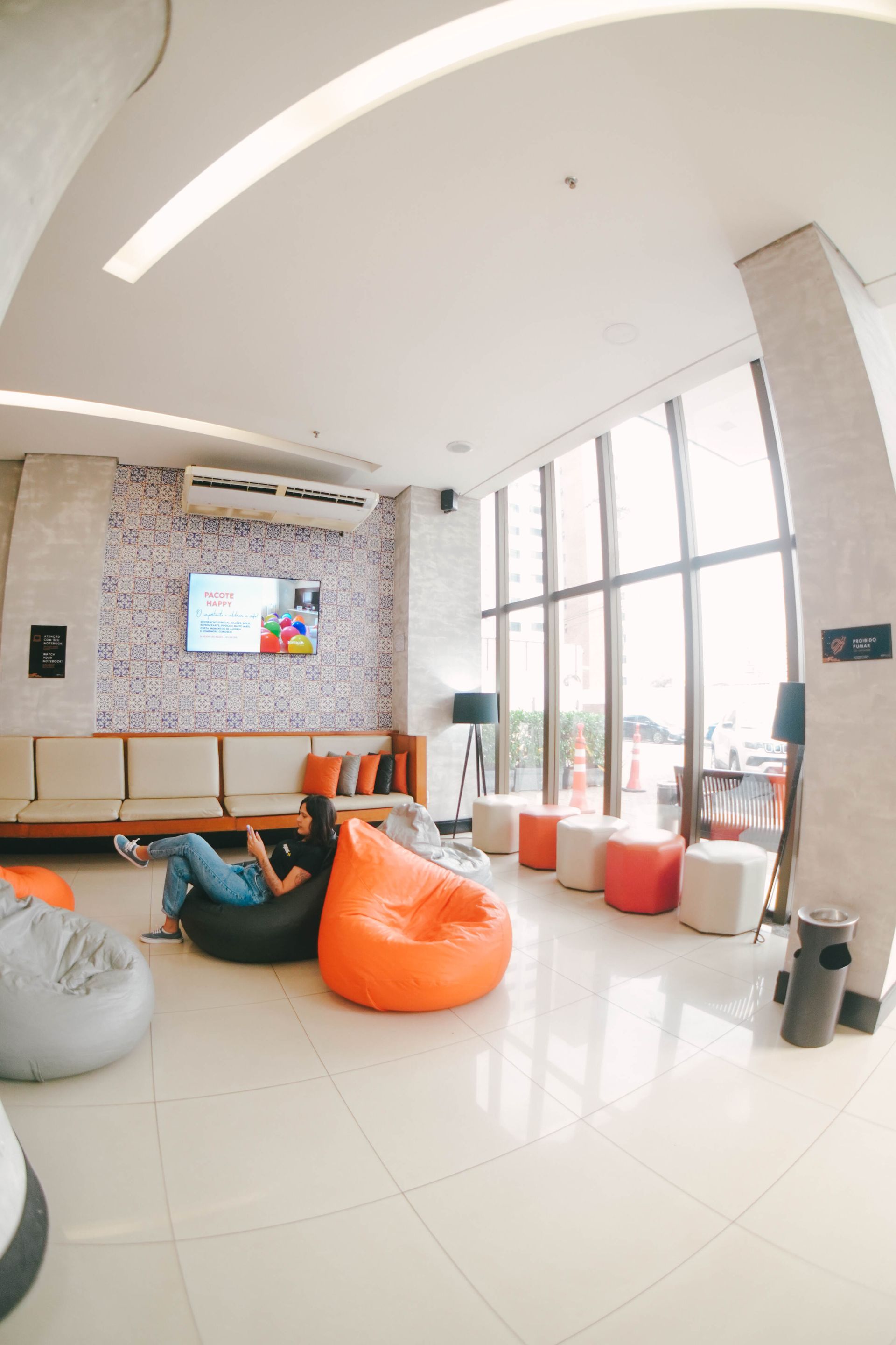 Sala de estar moderna com pufes, TV e grandes janelas. Alguém relaxa em um pufe.