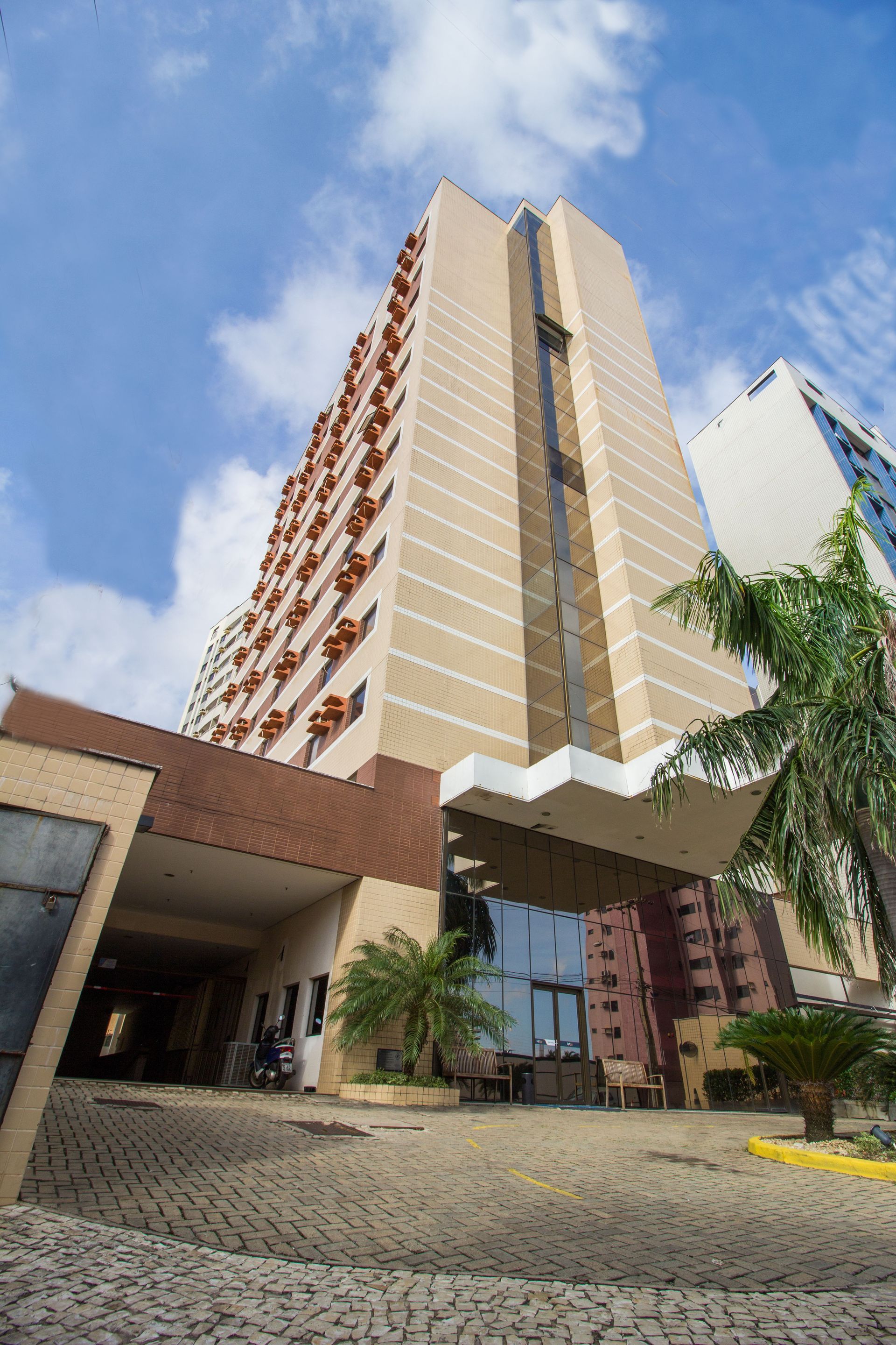 Prédio alto de hotel em tons de bege e marrom sob um céu azul, com palmeiras em primeiro plano.