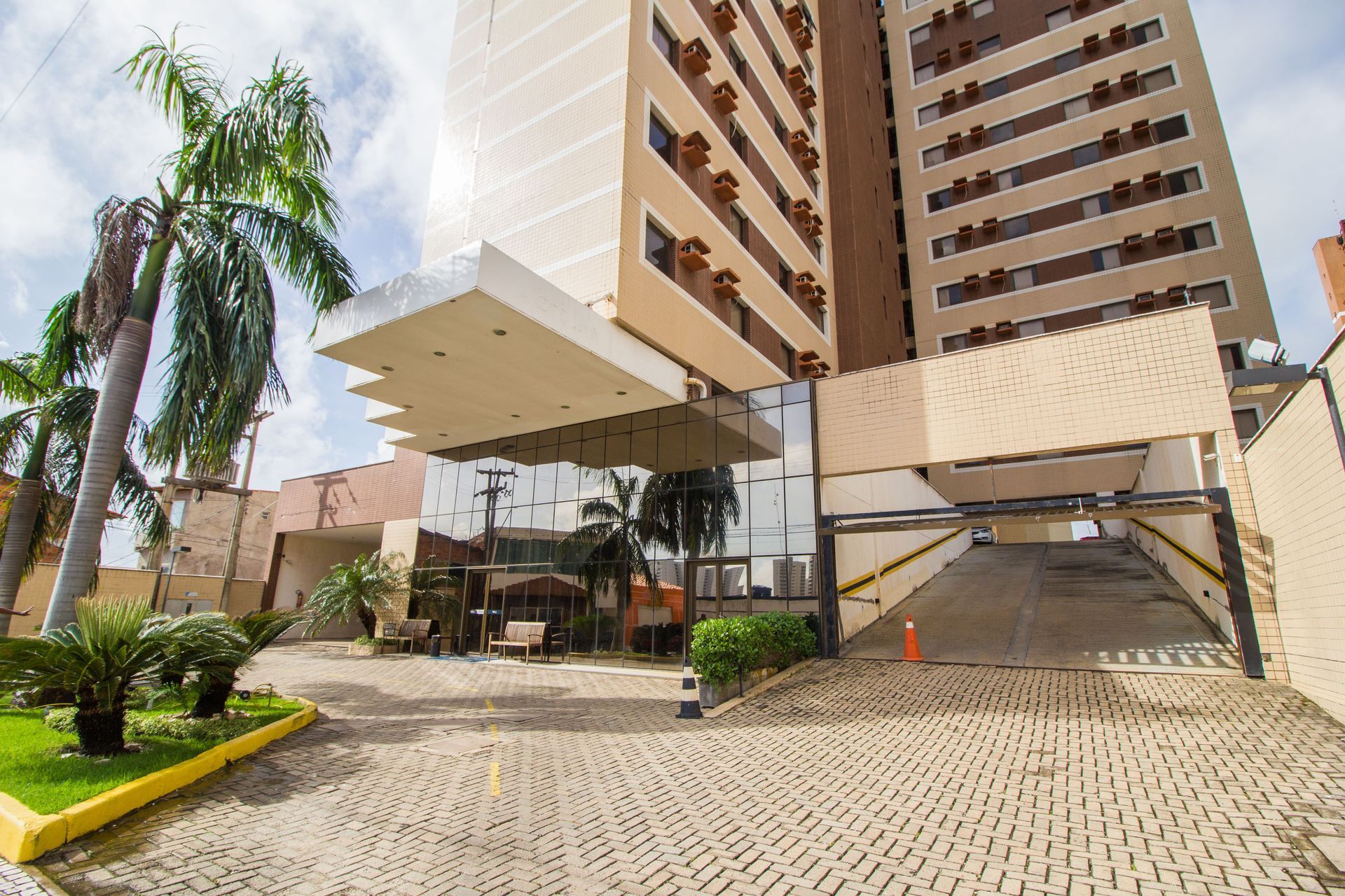 Prédio alto bege com entrada, estacionamento e paisagismo sob um céu nublado.