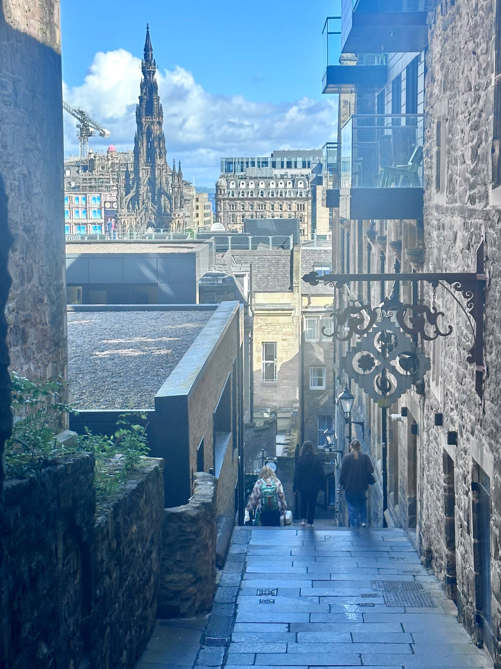 Narrow stone alley leading downhill to a historic city square with spires and old buildings in the distance