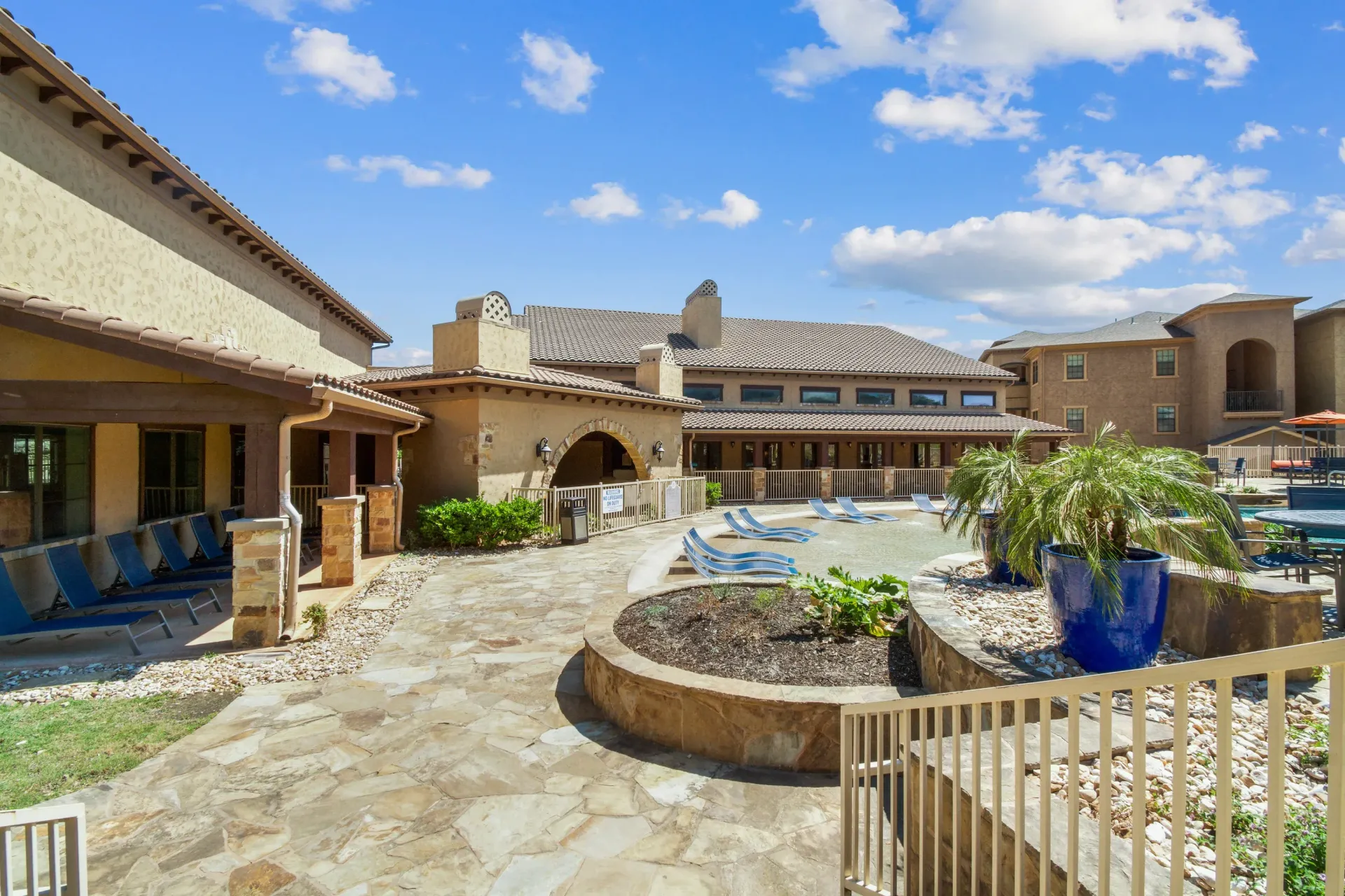 Swimming pool with a stone-covered gazebo, overlooking hillside and a building under a blue sky.