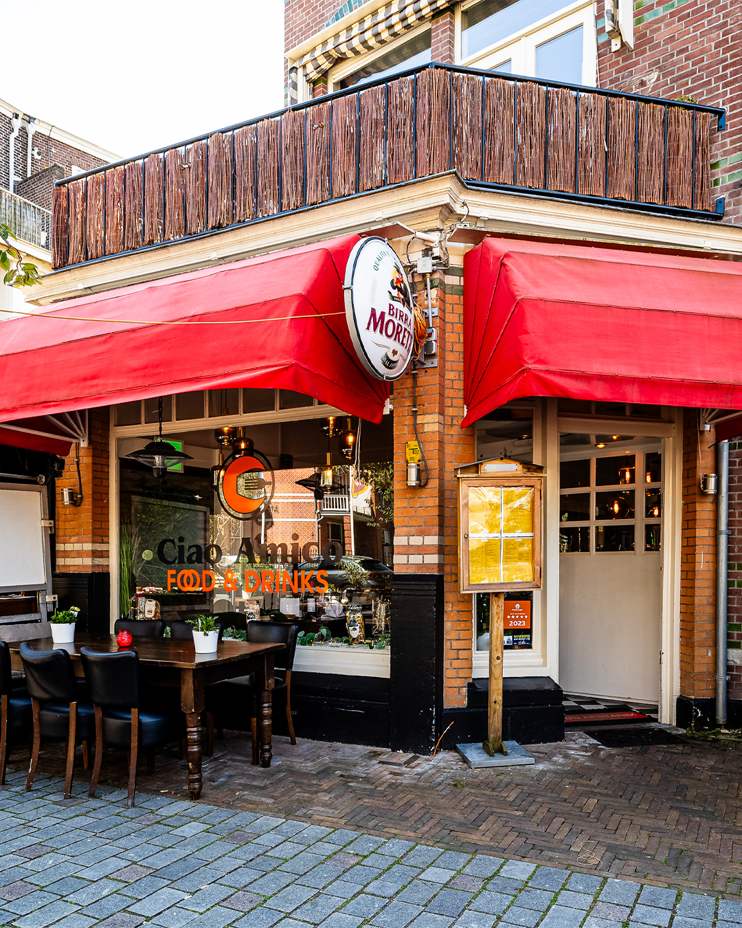Hoekrestaurant met rode luifel, uithangbord en terras. Bakstenen gebouw met grote ramen.