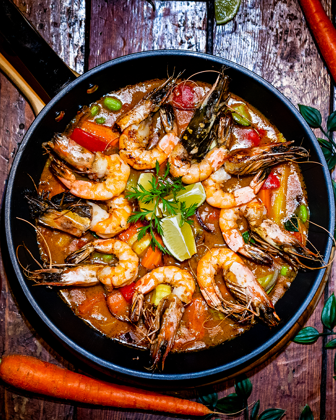 Garnalencurry met groenten in een zwarte pan, gegarneerd met limoen en kruiden. Wortelen zijn in de buurt te zien.
