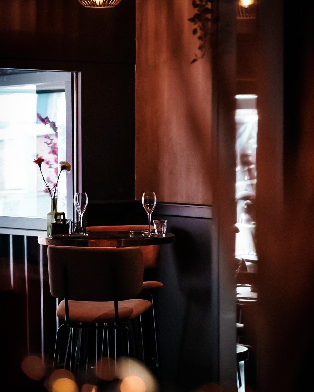 Een klein tafeltje met twee glazen en een vaas met bloemen, in een schemerig verlicht restaurant.