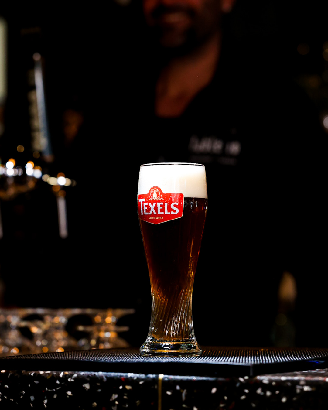 Een glas Texels-bier op de bar, met een wazige barman op de achtergrond.