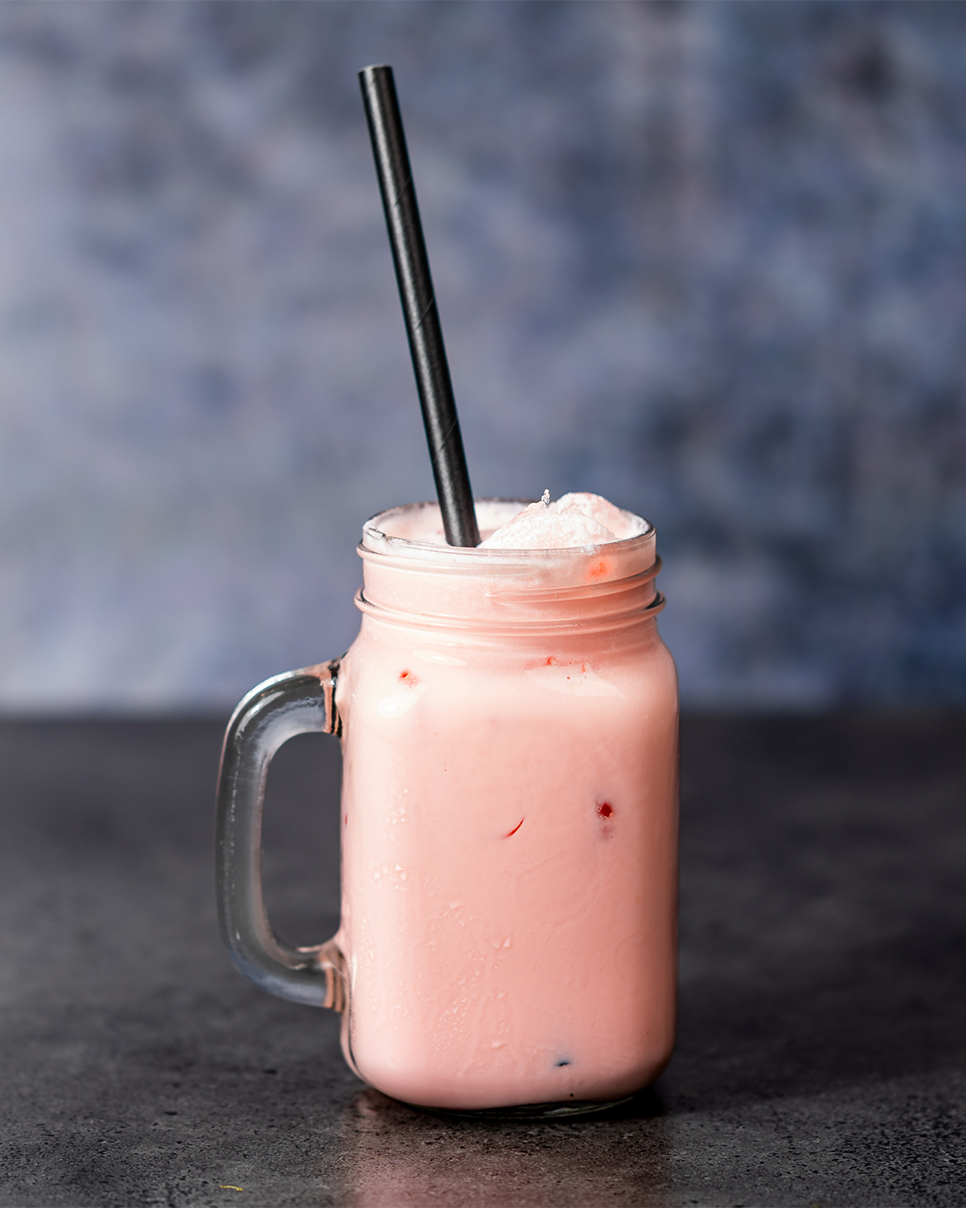 Roze drankje in een glazen pot met een zwart rietje, geplaatst op een donkere ondergrond.