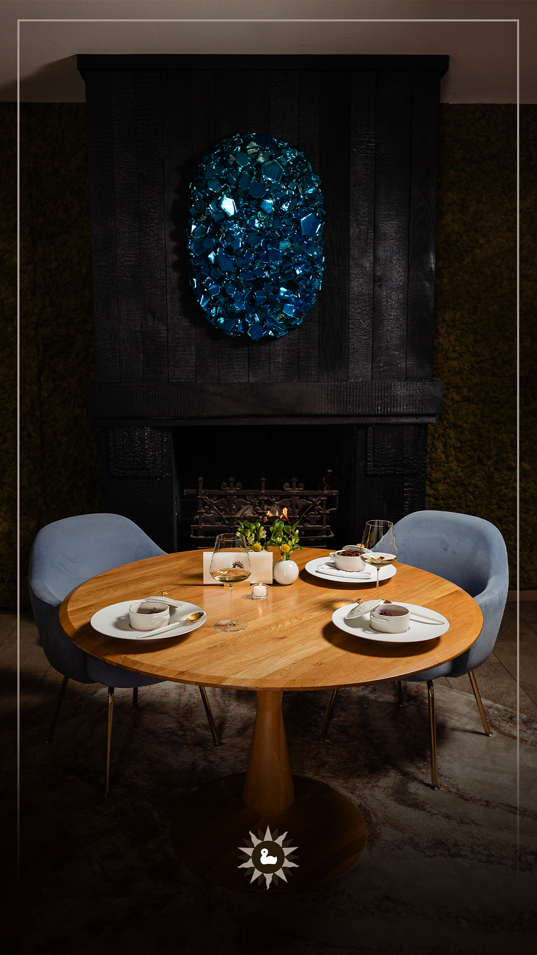 Gezellige eetscène: ronde houten tafel gedekt voor drie personen, met blauwe stoelen. Donkere open haard met blauwe geode-decoratie.