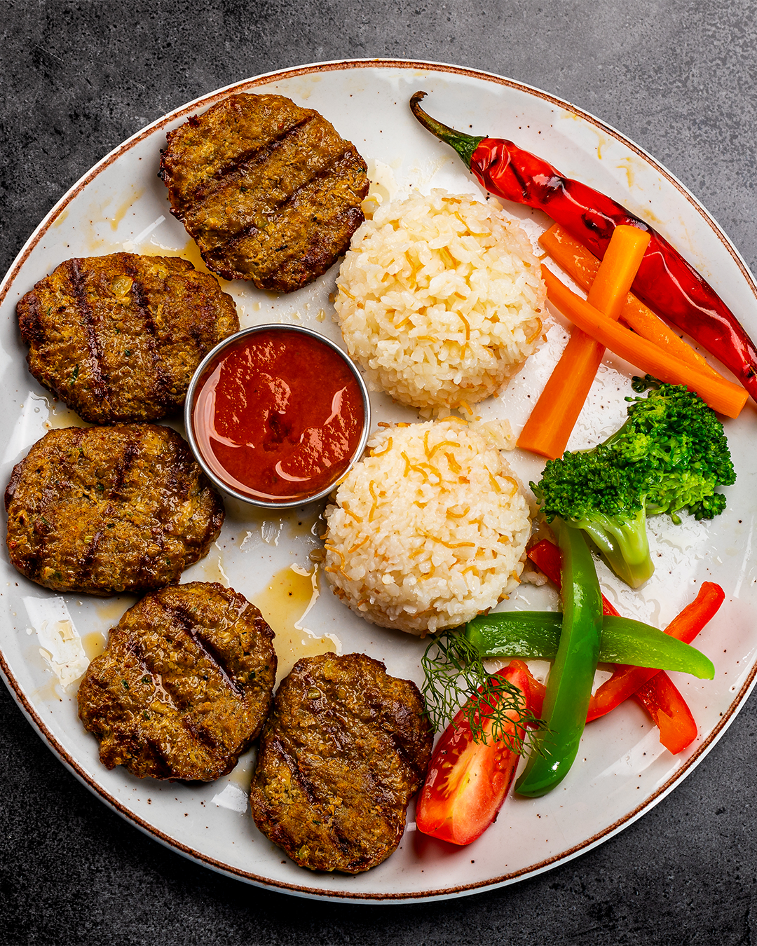 Een bord met gegrilde burgers, rijstballetjes, saus en groenten zoals wortels, broccoli en paprika's.
