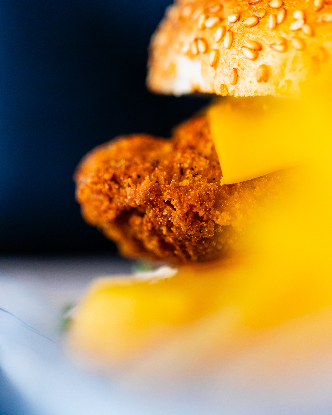 Close-up van een sesamzaadbroodje met een gefrituurde kipburger en kaas, tegen een donkere achtergrond.