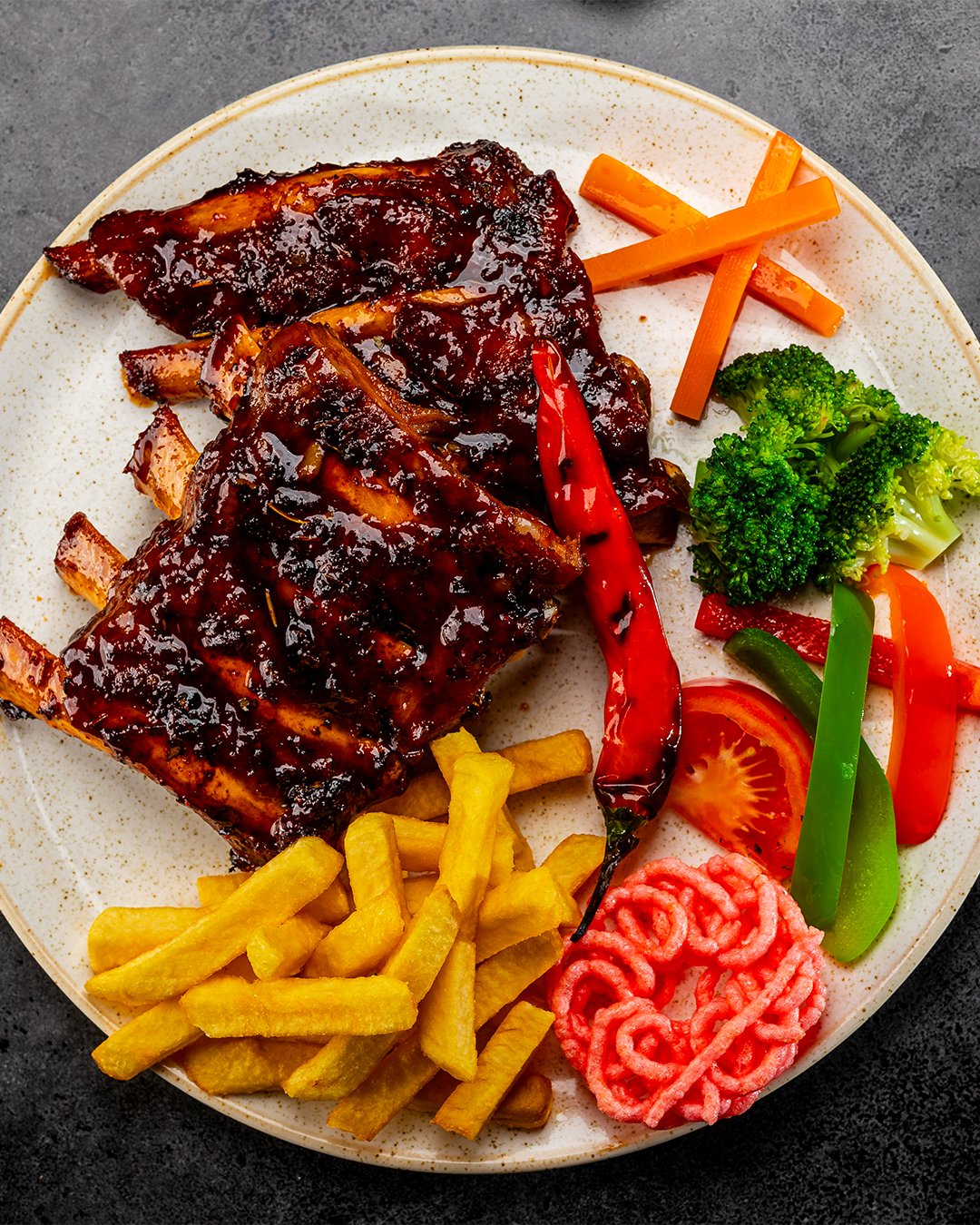 Een bord met geglazuurde ribbetjes, friet, groenten en een rode paprika.