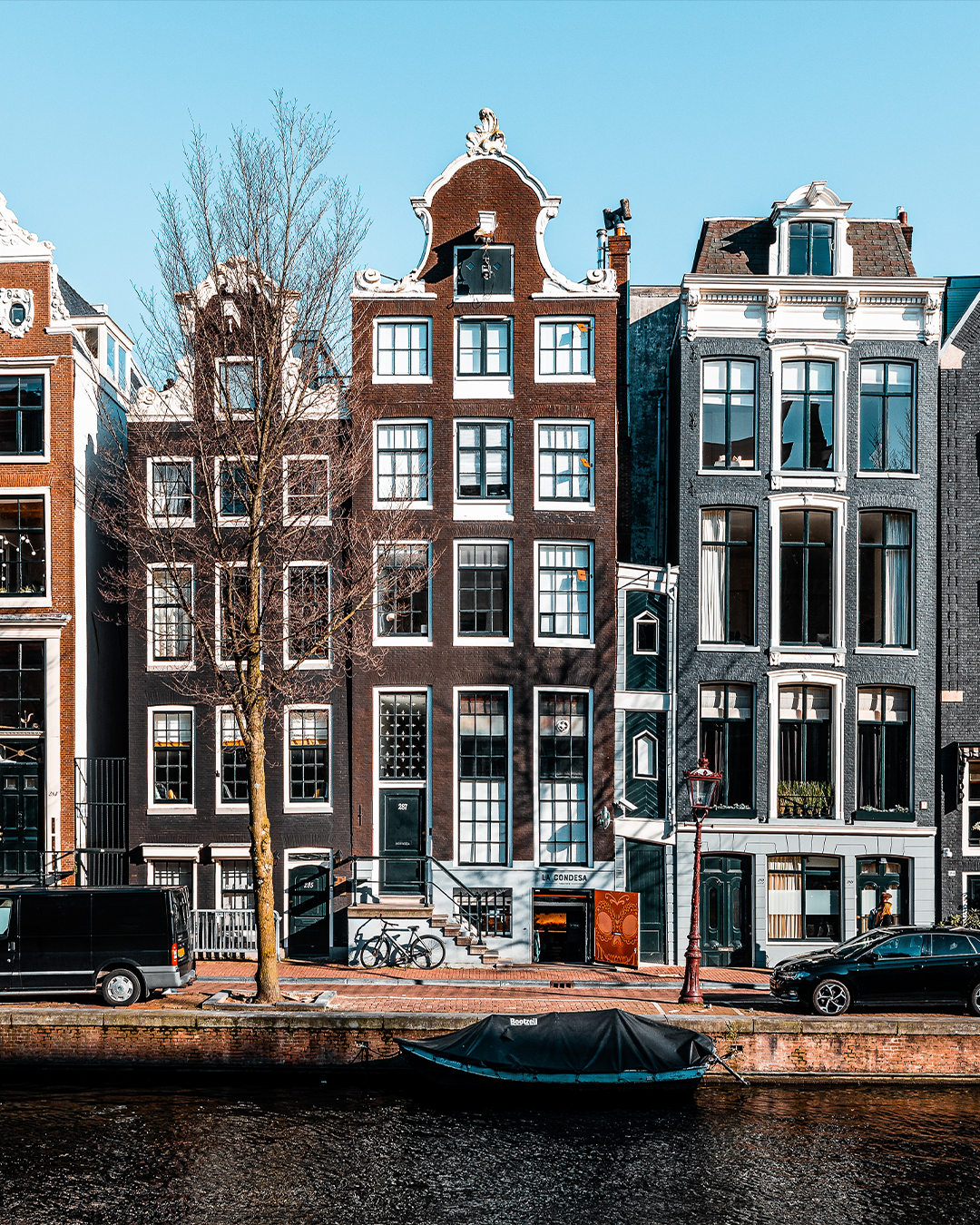 Amsterdamse grachtenpanden, bakstenen gevels, boot in het water, geparkeerde auto's, heldere hemel.