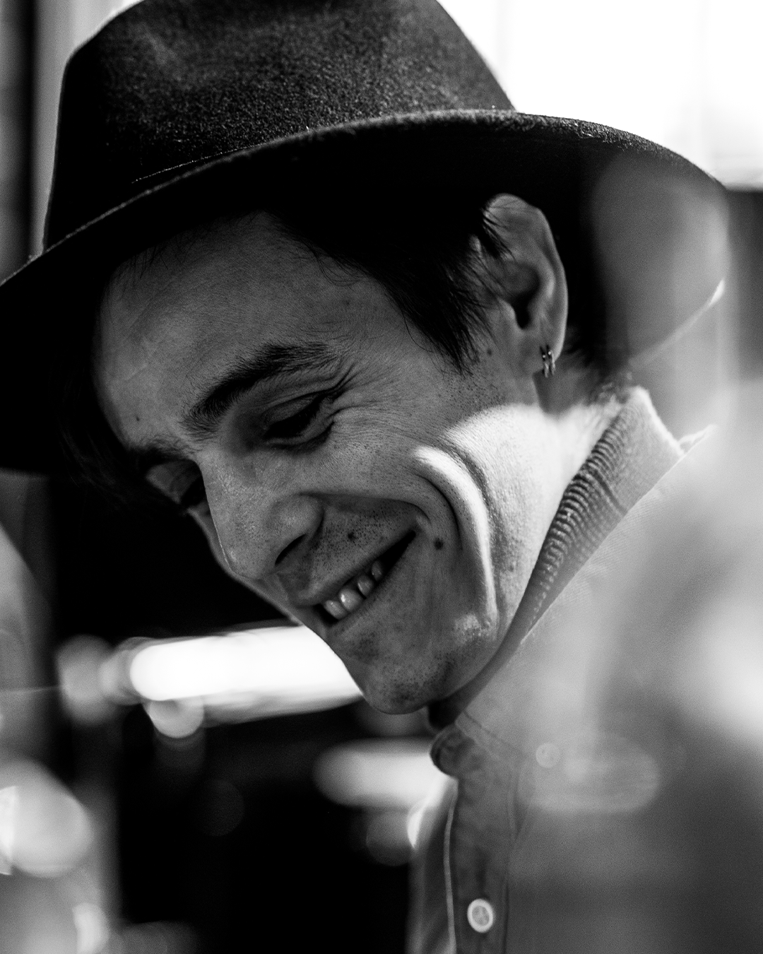 Man met fedora glimlacht en kijkt naar beneden; zwart-witfoto.
